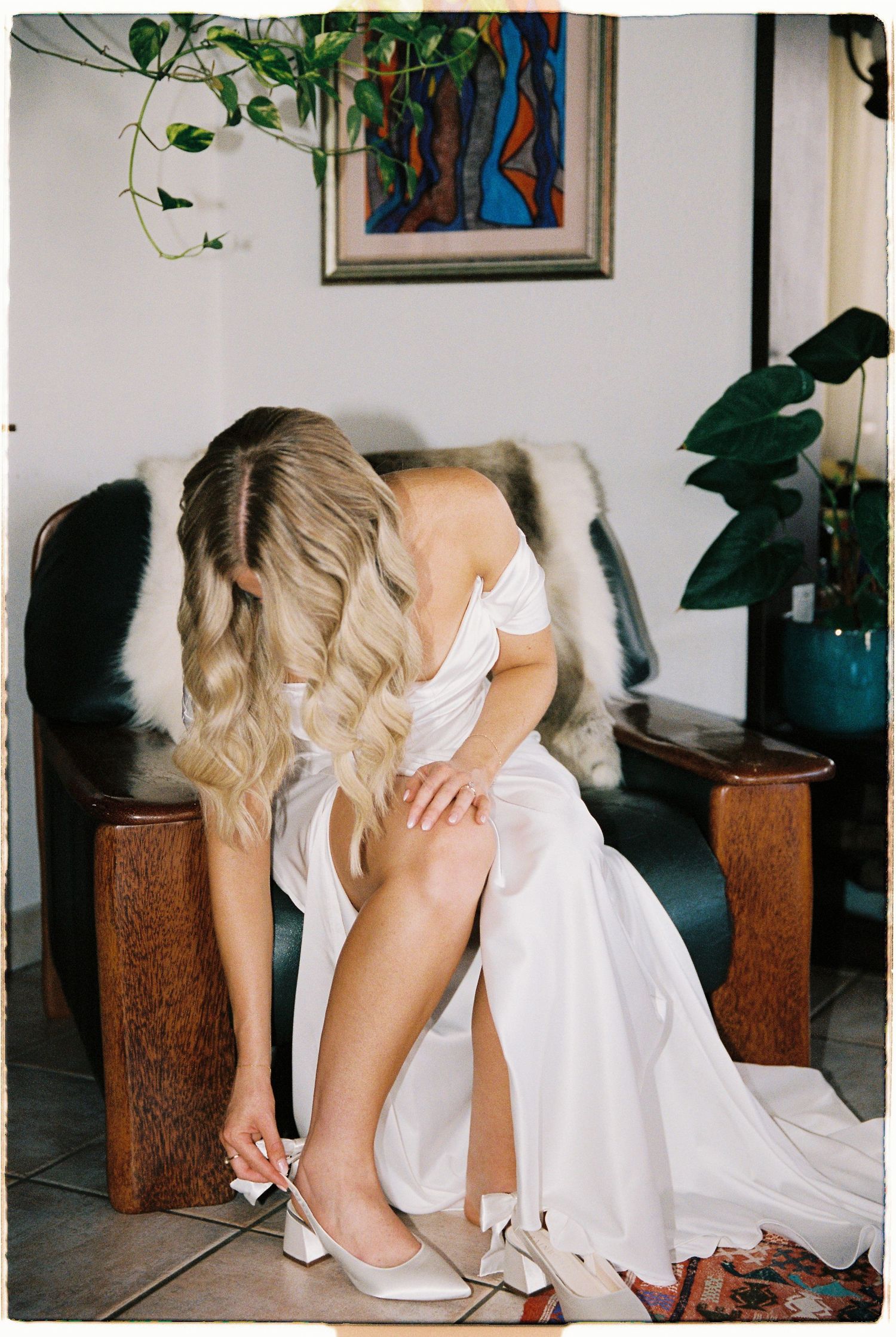 Blonde bride in white off-shoulder wedding dress putting on heels while seated in vintage leather chair indoors.