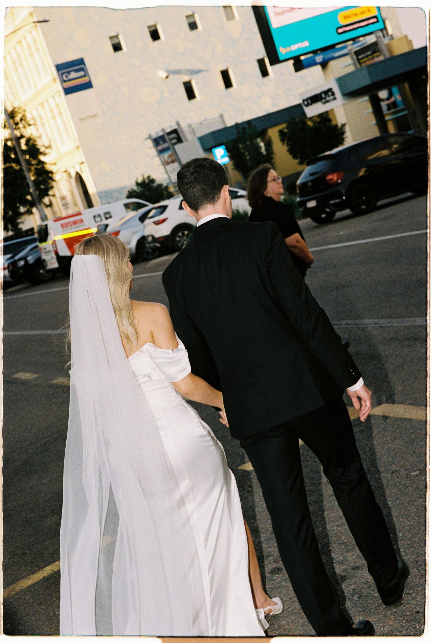 Bride in white gown and veil walks with groom in black suit across urban street, third person beside them.