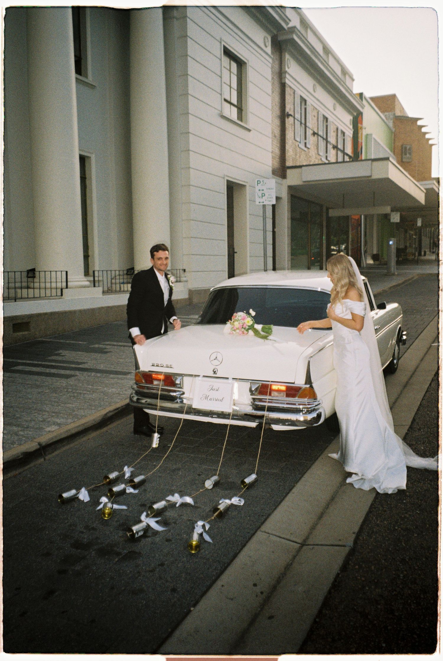 Bride and groom pose beside a vintage white Mercedes-Benz wedding car decorated with flowers and trailing ribbons on a city street.
