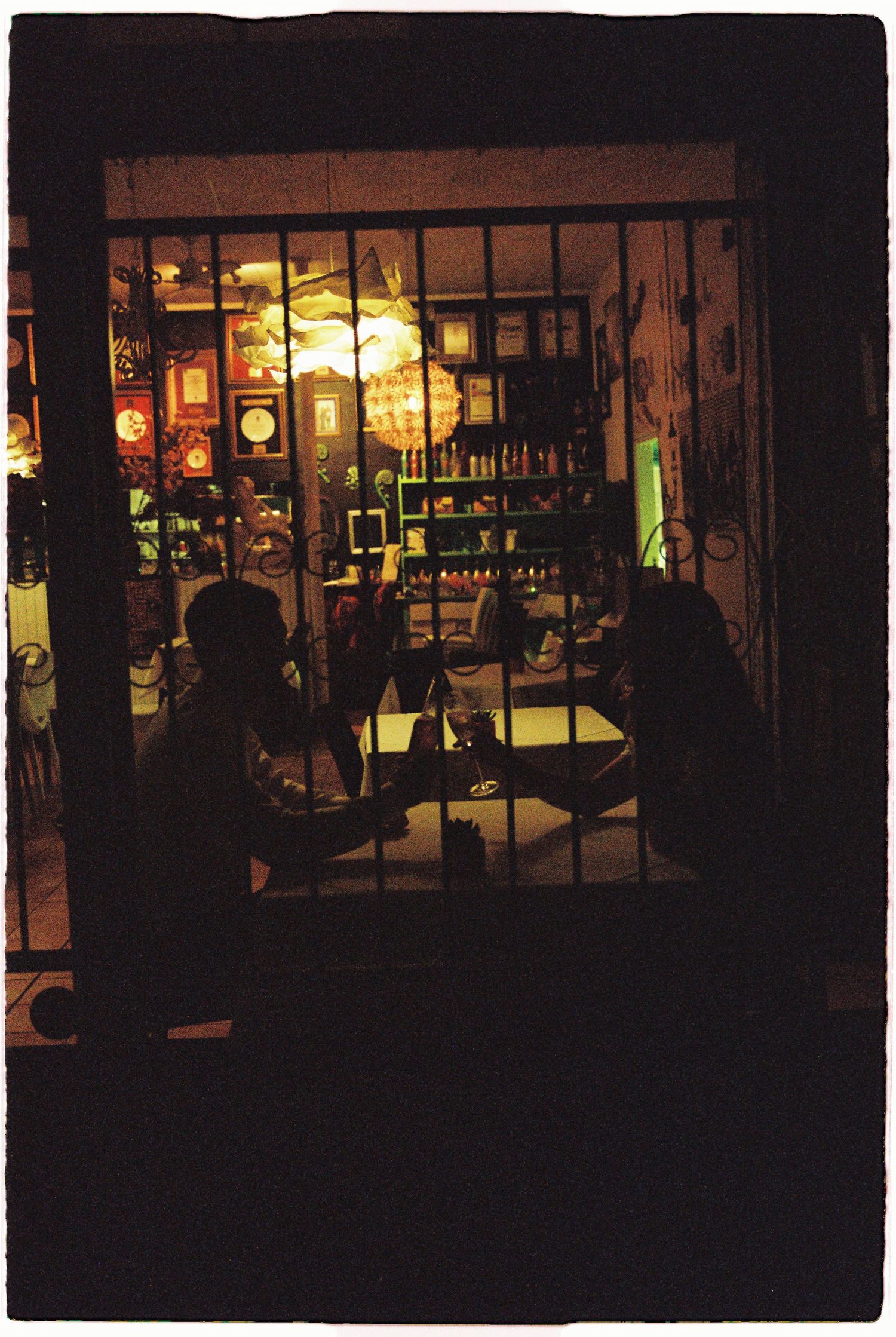 Silhouetted people sitting at a table inside a warmly lit bar with shelves of bottles behind bars.
