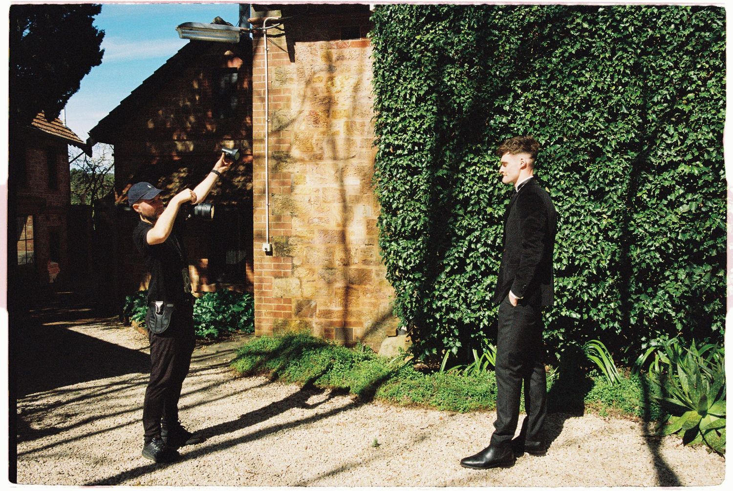 Photographer captures groom in black suit standing outdoors beside rustic wooden door and green ivy wall.