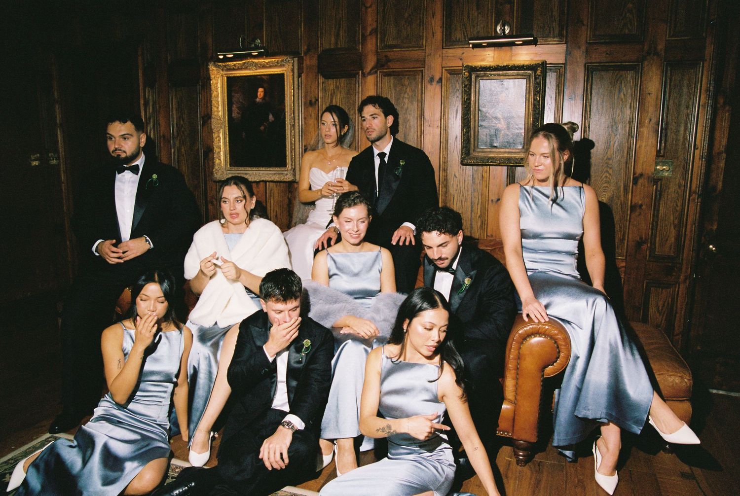Wedding party group photo in wood-paneled room wearing grey bridesmaid dresses and tuxedos.