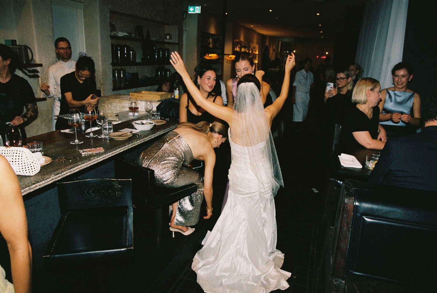 Bride in white wedding gown with veil dancing joyfully with guests at a lively wedding reception bar.
