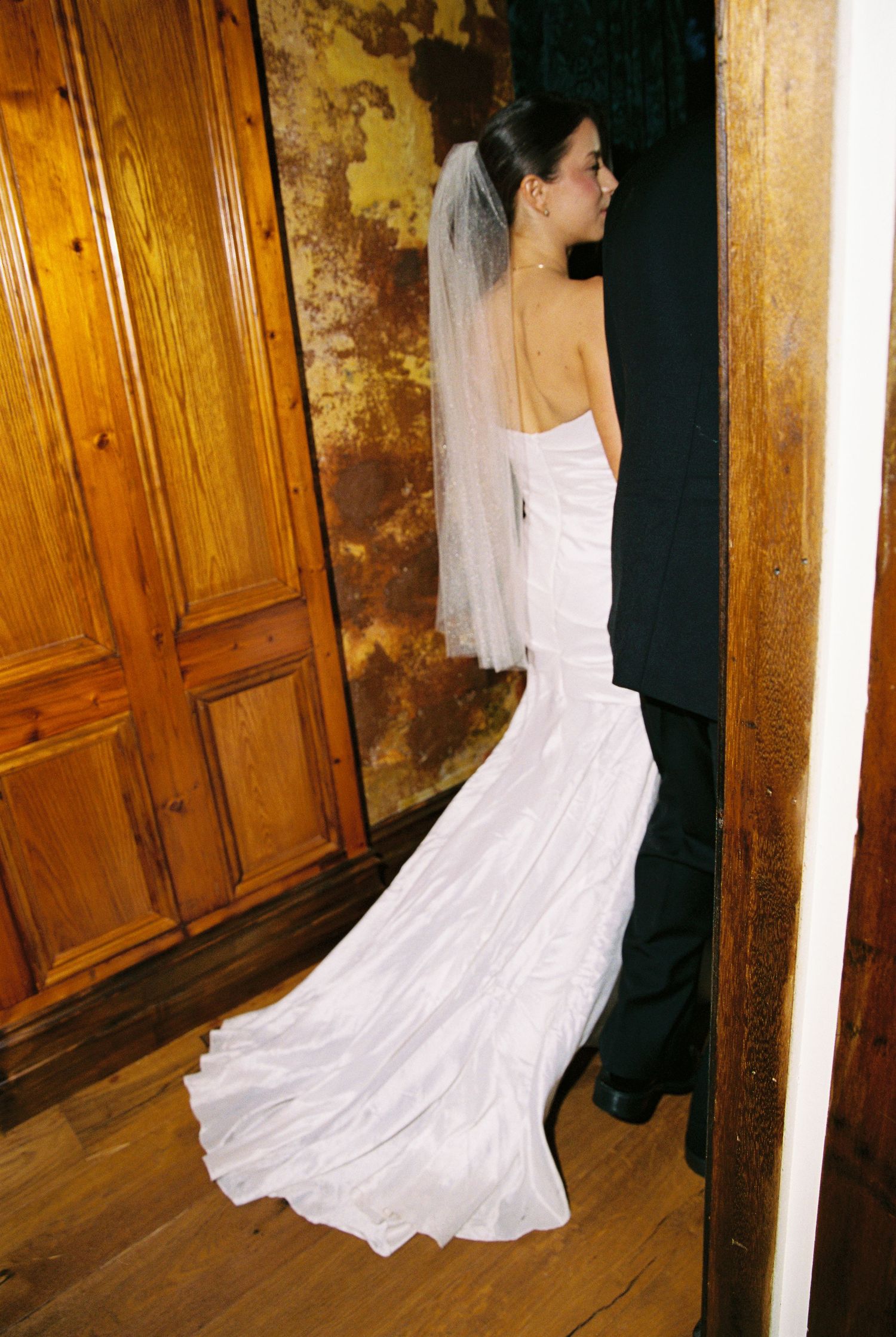 Bride in a sleeveless mermaid wedding gown with veil standing in a wooden doorway from behind.