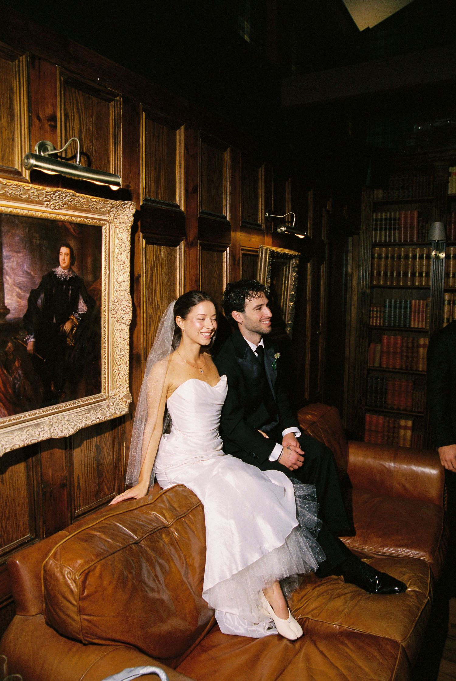 Bride and groom sitting together on a leather sofa in an elegant wood-paneled library with vintage portraits.