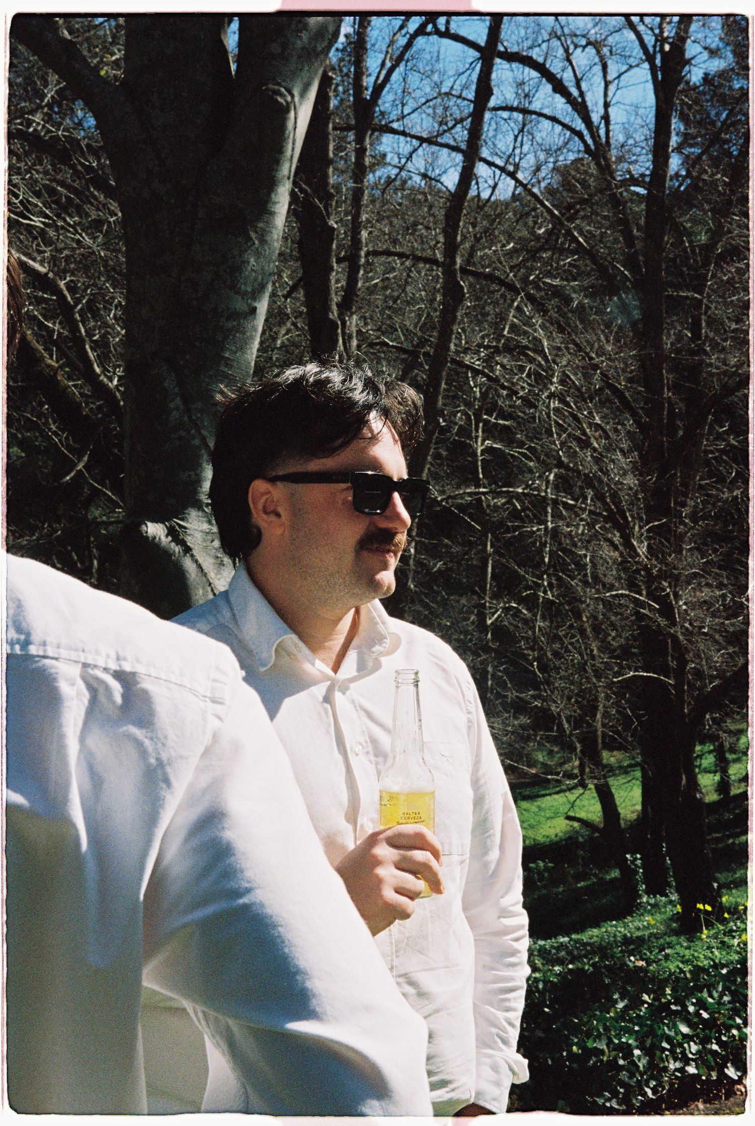 Man in white shirt and sunglasses holding a beer bottle outdoors at a wedding.