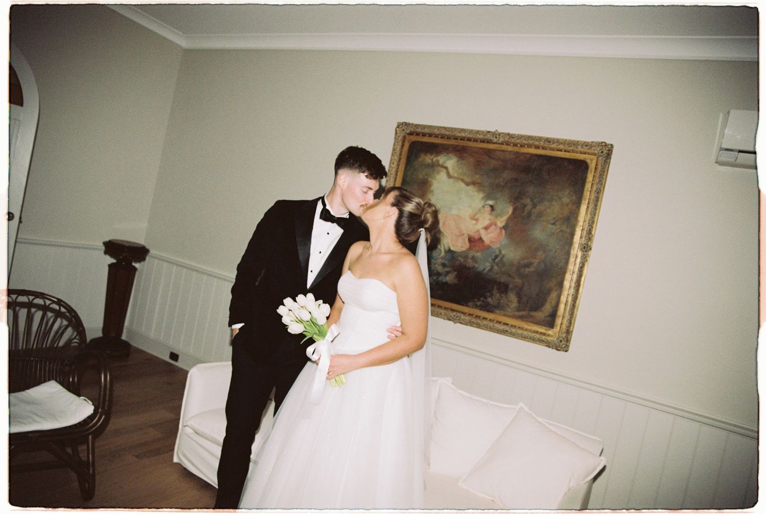 Bride and groom sharing a kiss indoors beside a classic oil painting on wall.