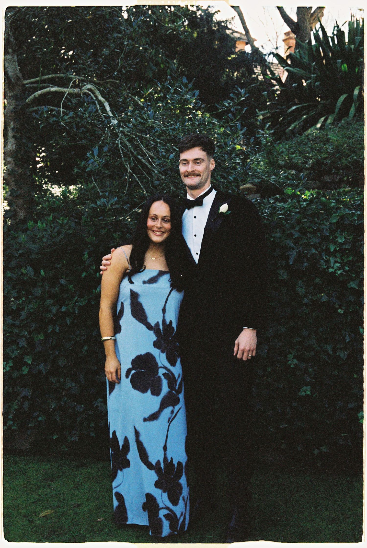 Couple posing together in formal attire in a garden, man in tuxedo and woman in blue floral gown.