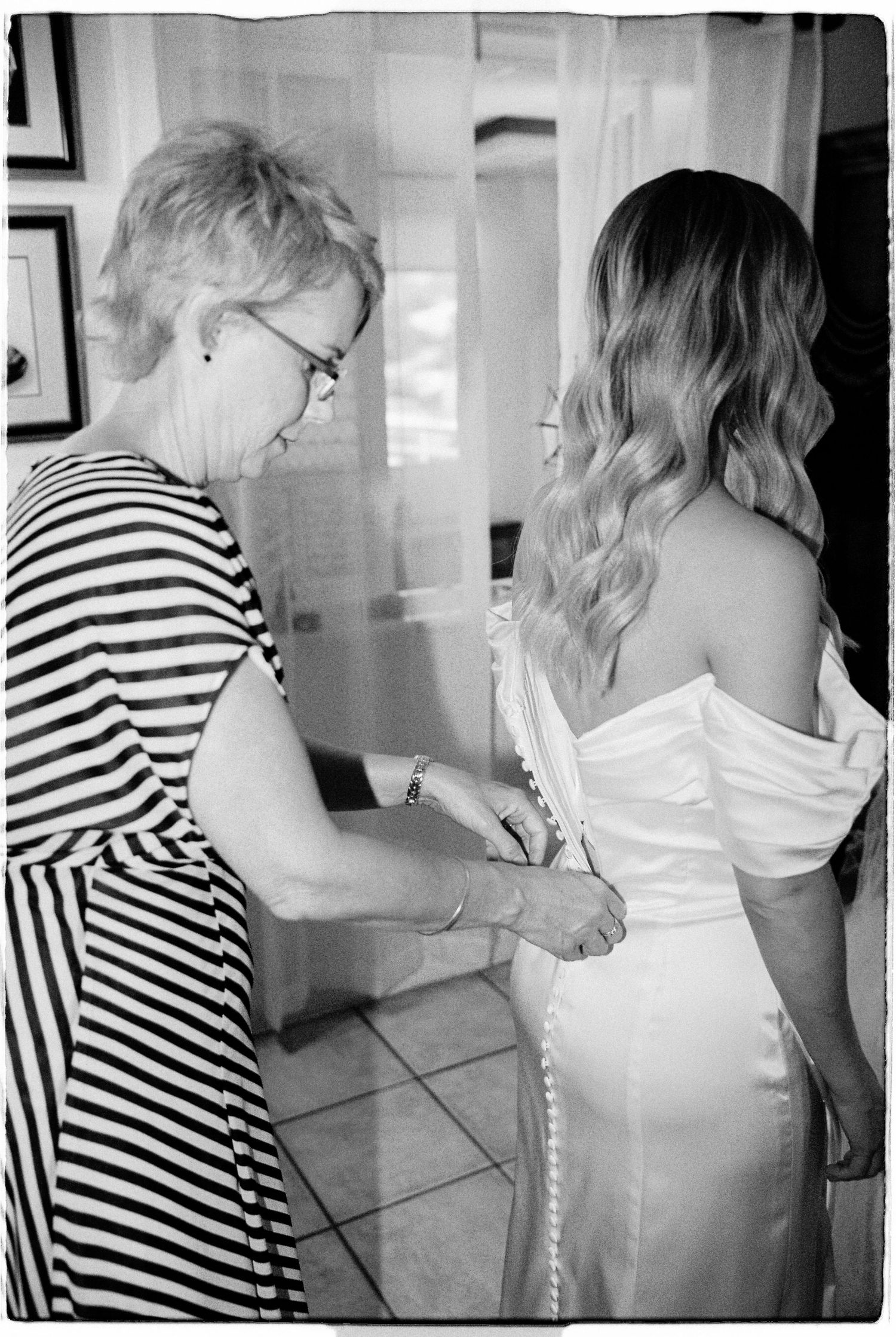 Older woman helping a bride button her white wedding dress in a black and white photo.