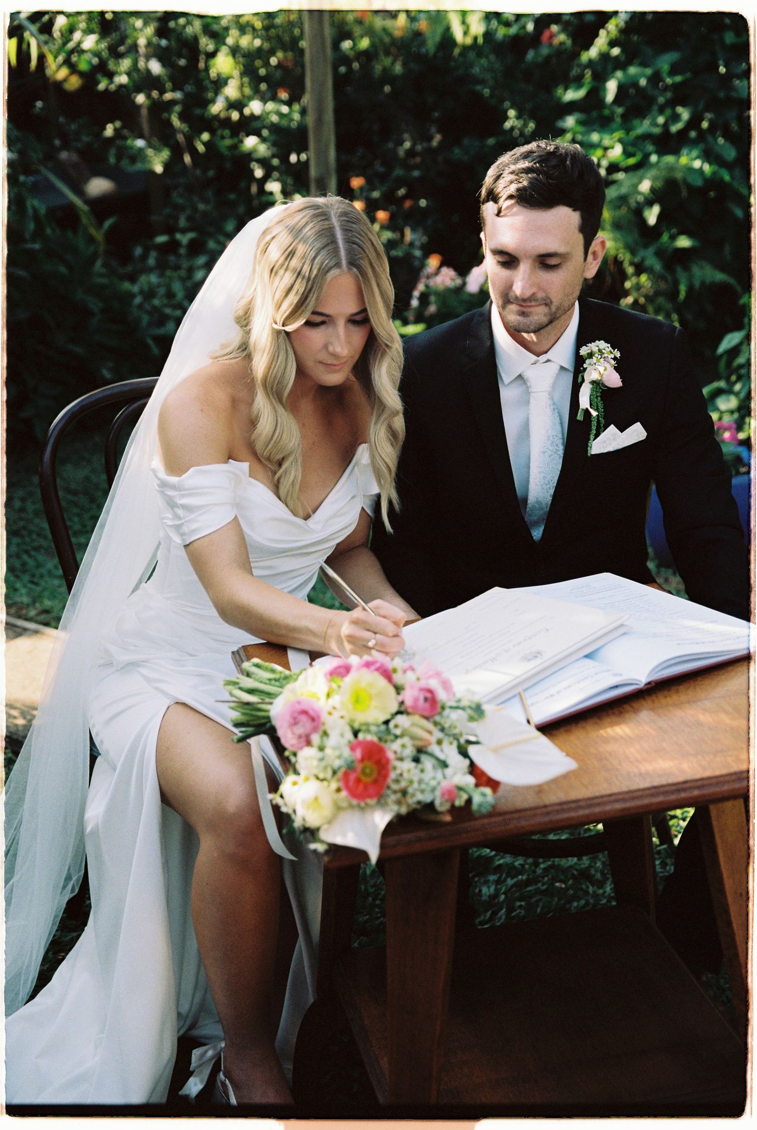 Bride in white gown signs wedding register beside groom in black suit with floral bouquet on table.
