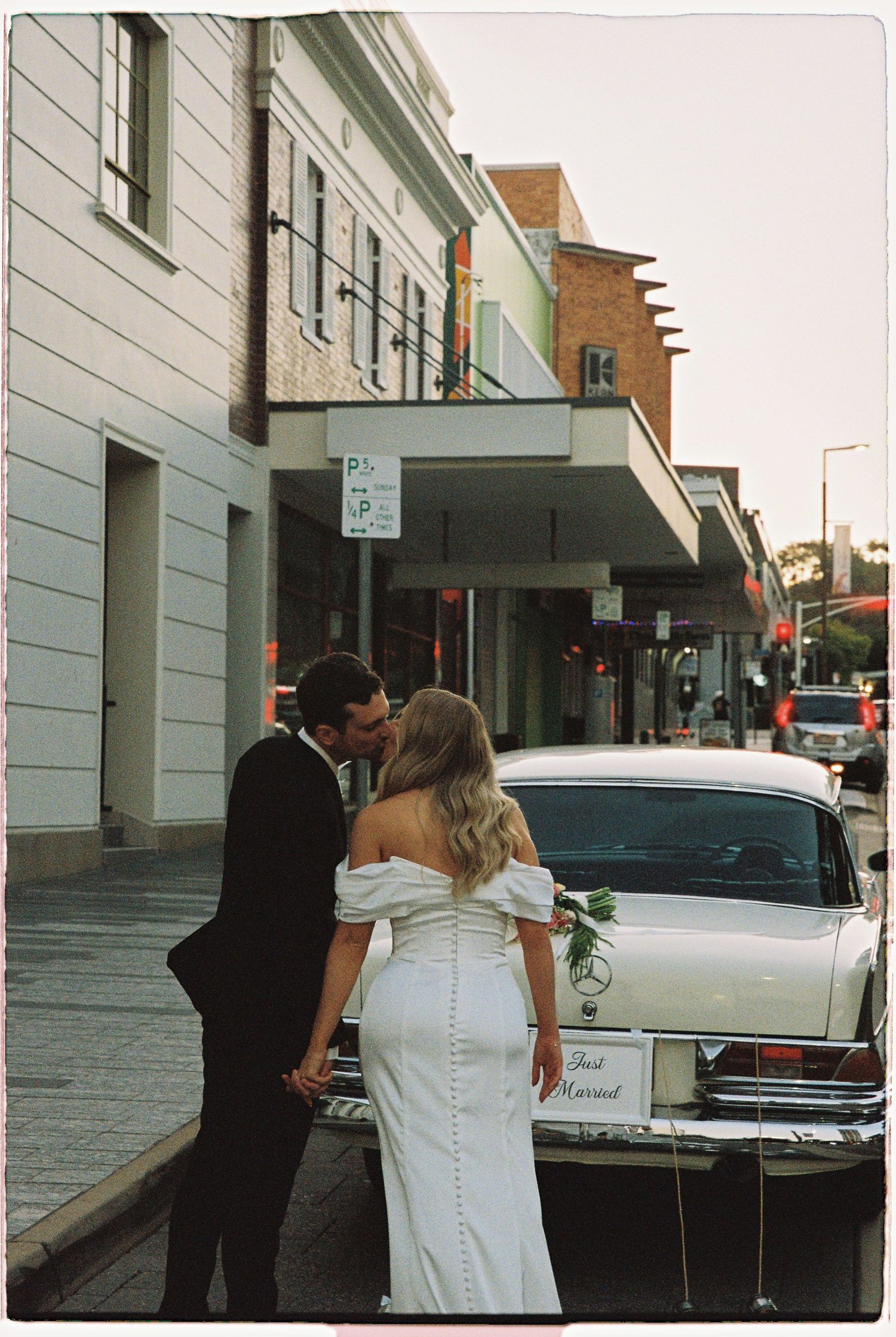 Newlyweds kissing beside a vintage white Mercedes wedding car on a city street at dusk.