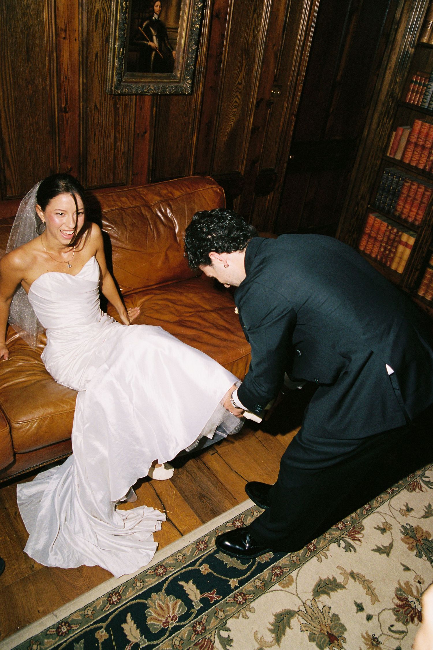 Groom kneeling to help bride with her shoes on a leather sofa in a library.