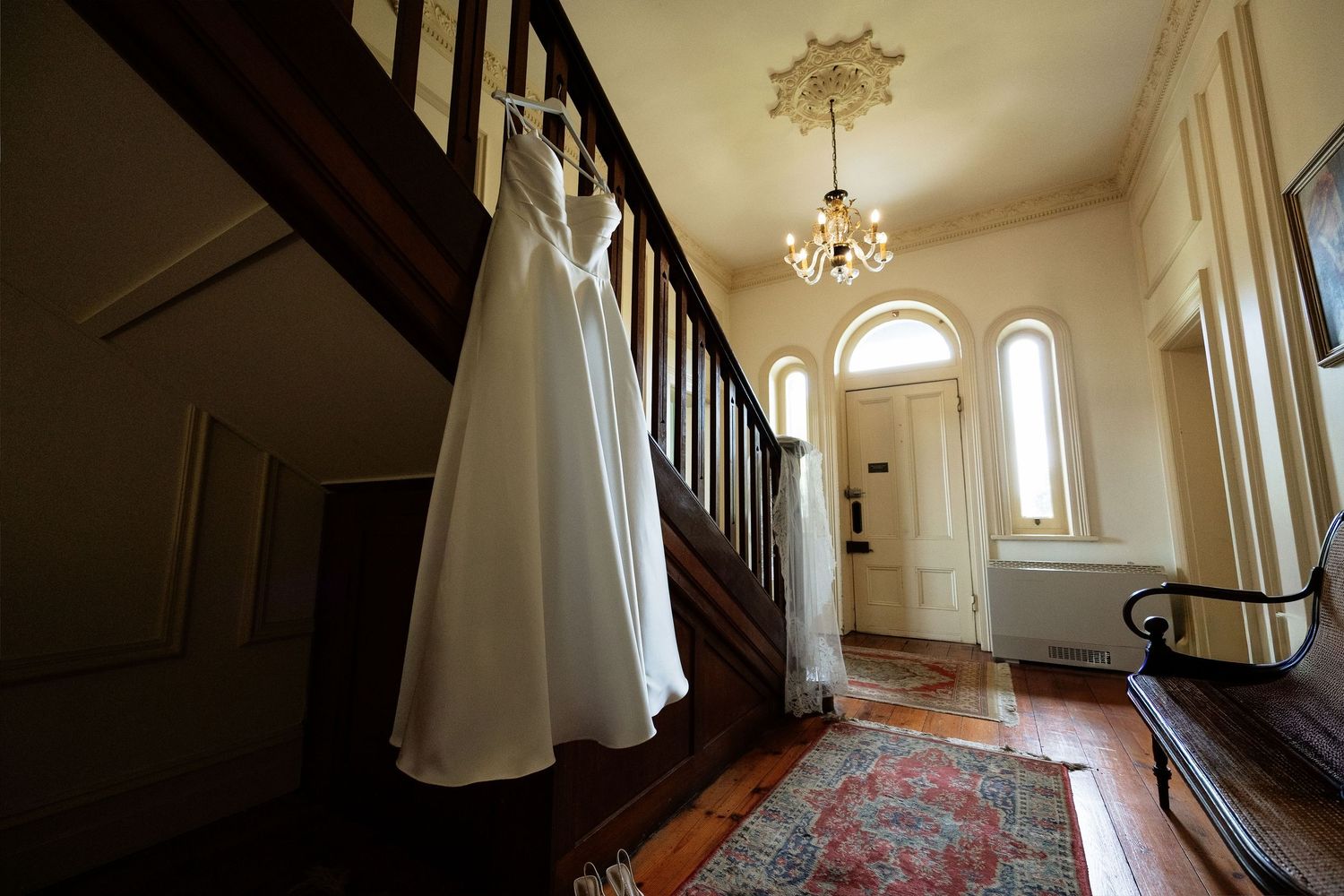 White wedding dress hanging on staircase banister in elegant foyer with chandelier and hardwood floors.
