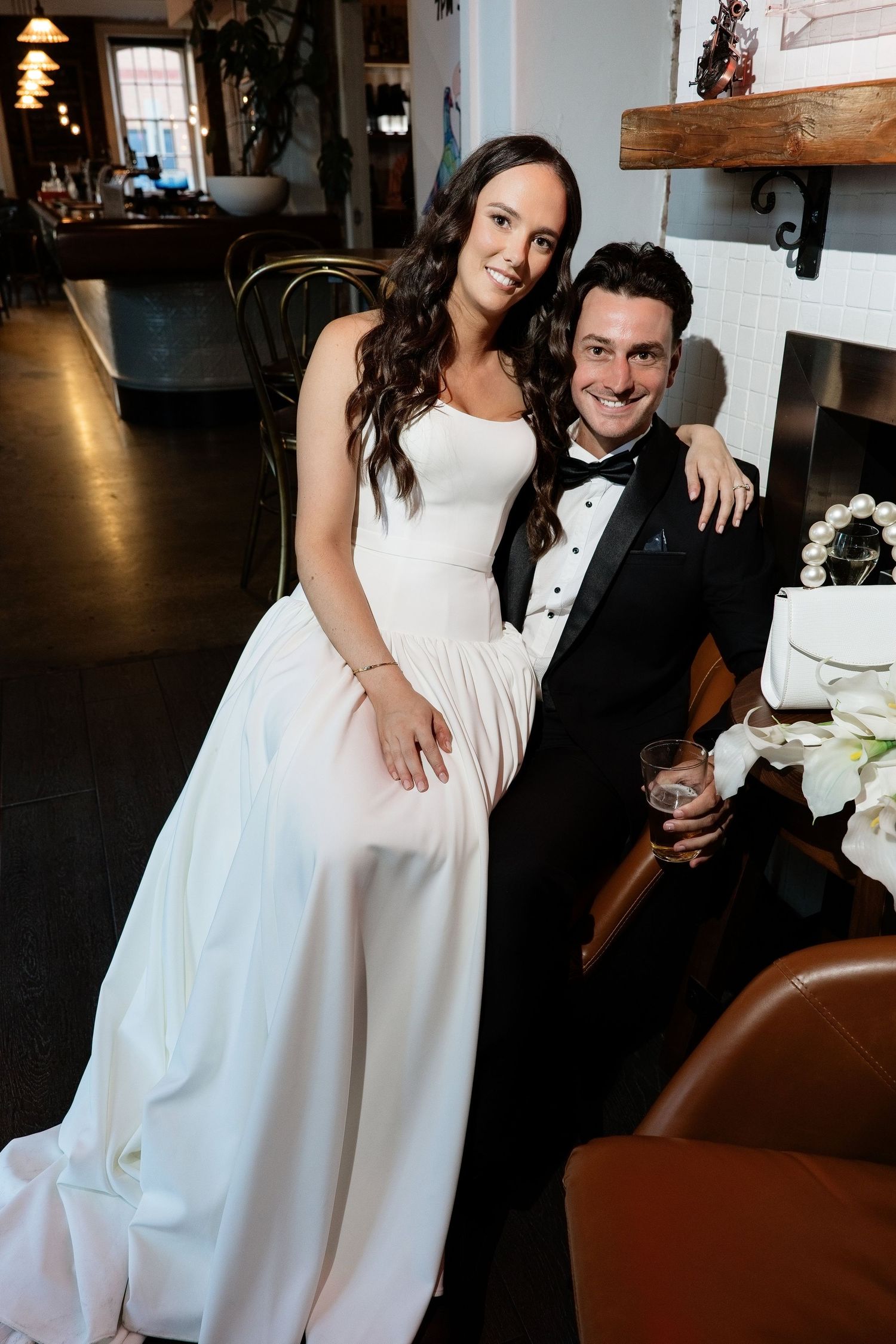 Smiling bride and groom posing together near a fireplace, bride in white gown, groom in black tuxedo.