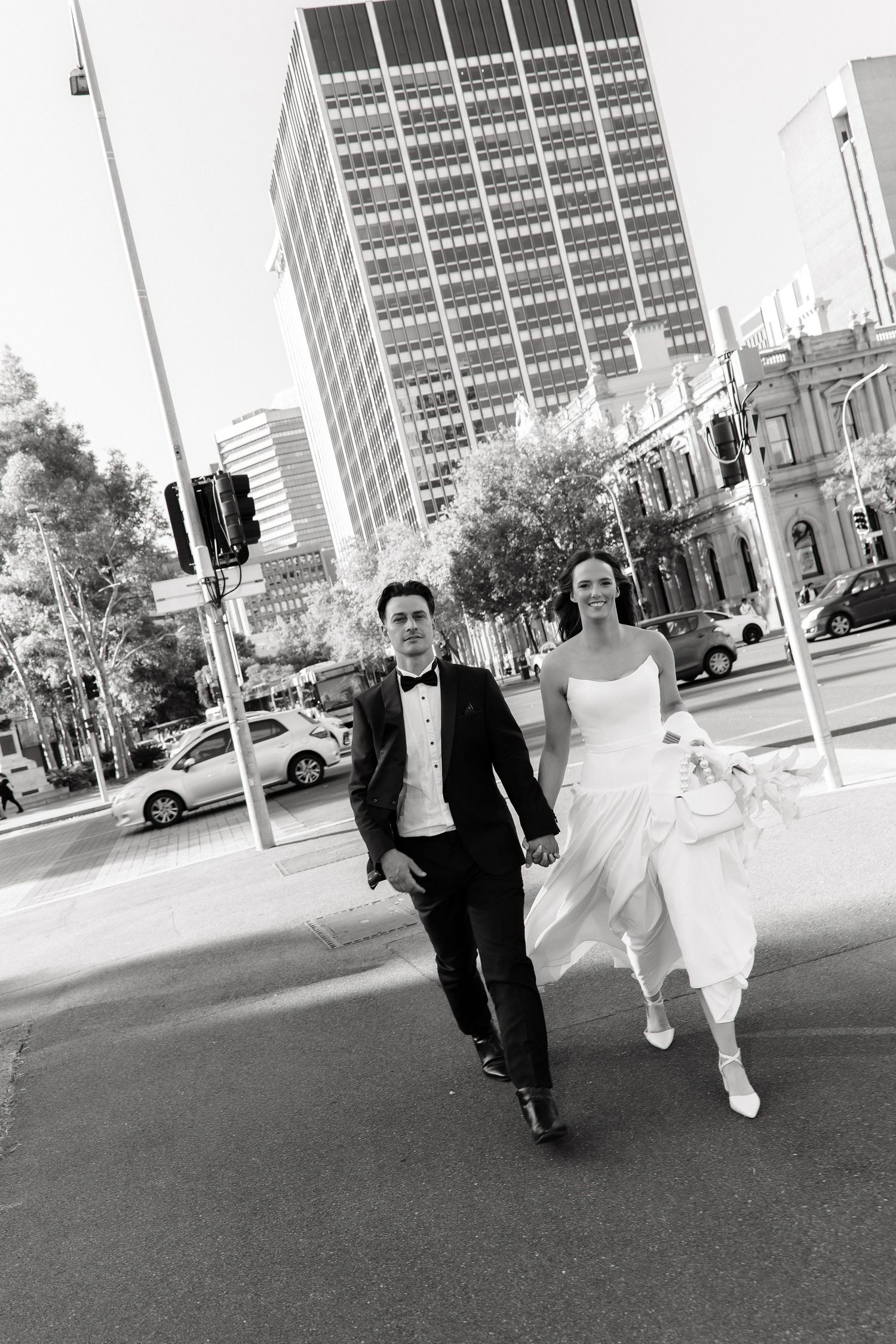 Smiling bride and groom hold hands while crossing a city intersection with tall buildings in the background.