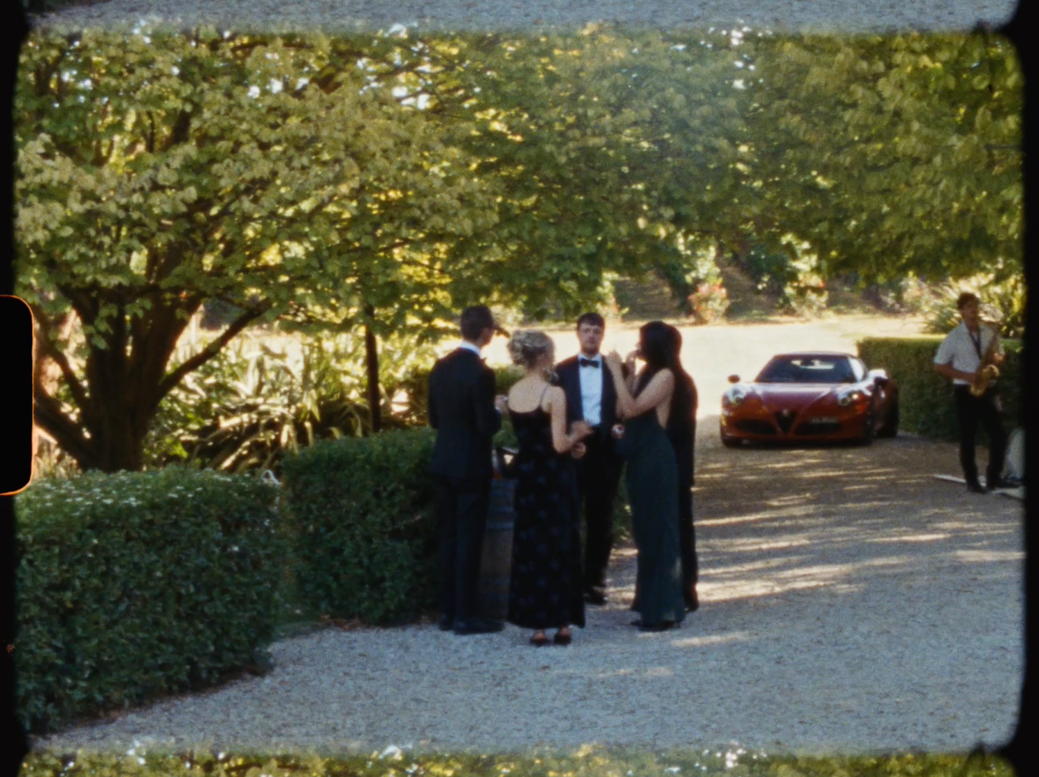 Elegantly dressed wedding guests socializing on a gravel path beside a red sports car and lush trees.