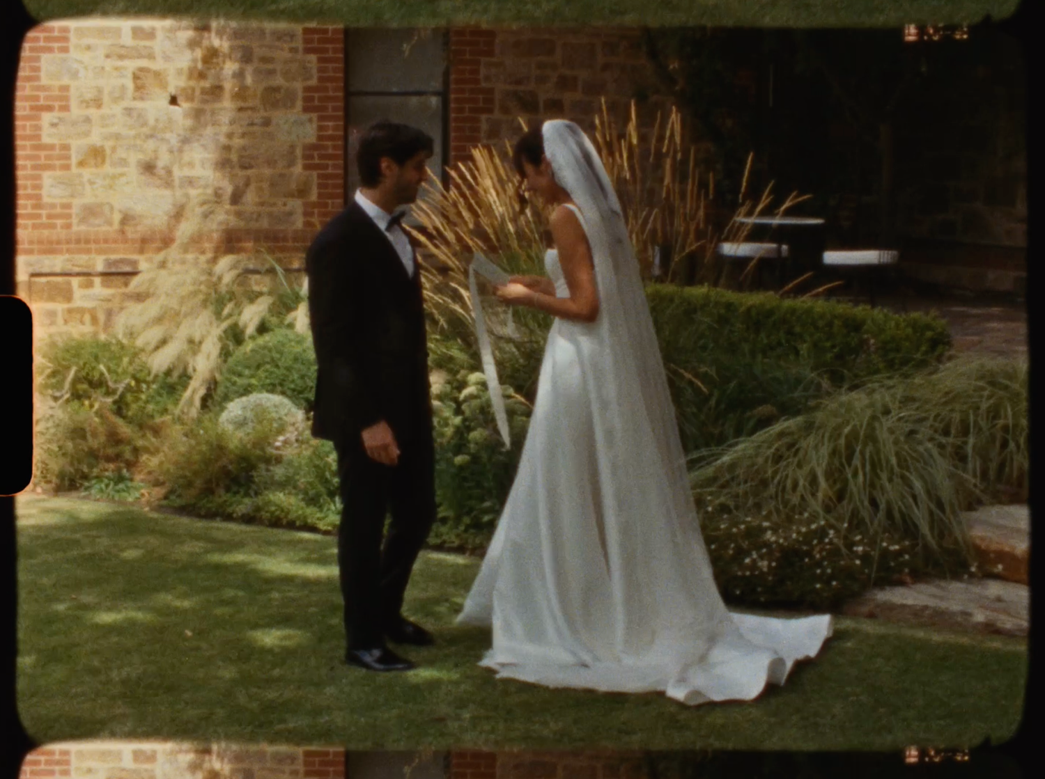 Bride in white gown with veil and groom in black tuxedo standing in garden at wedding.