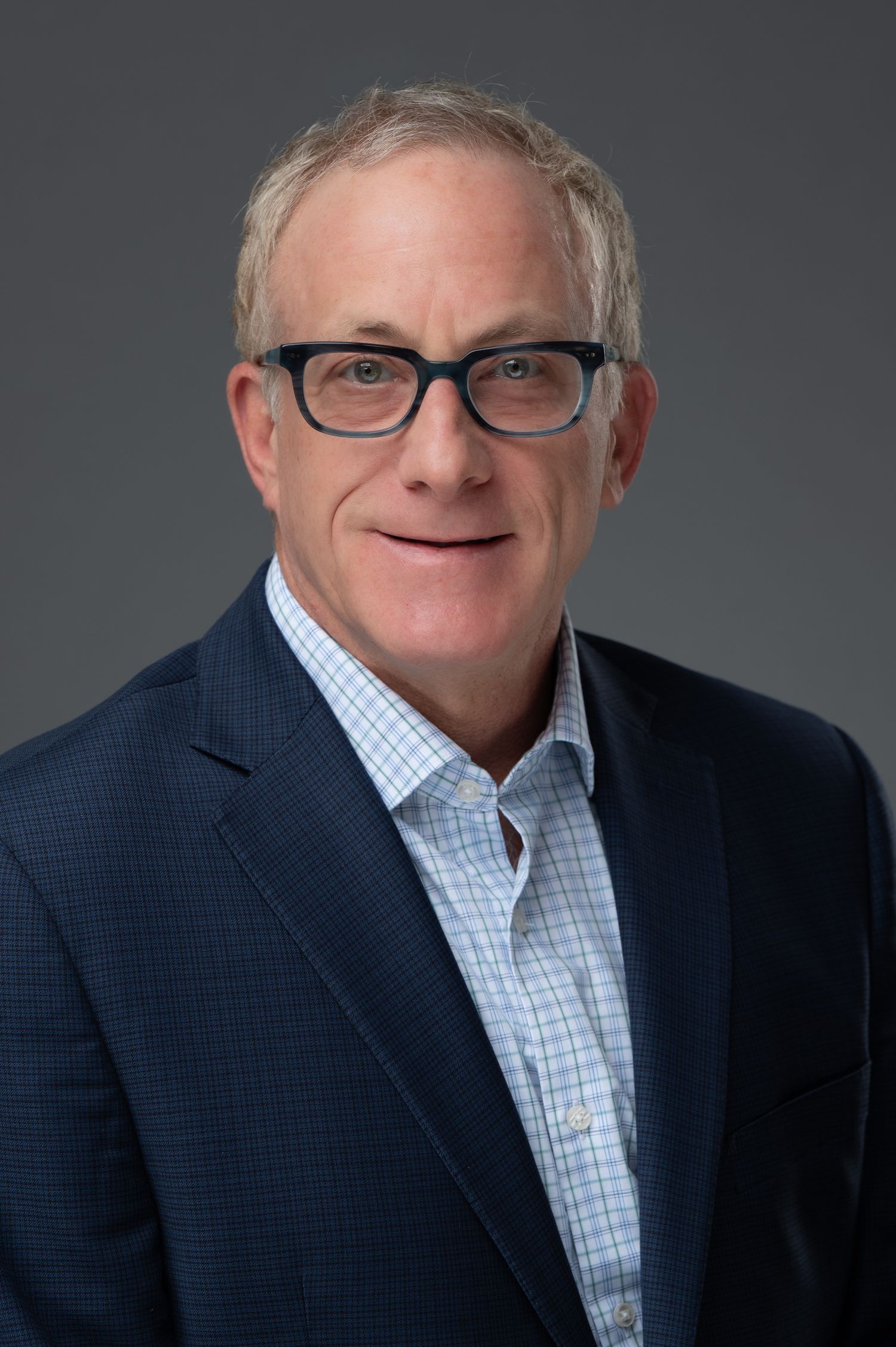 Professional headshot of a middle-aged man wearing dark-rimmed glasses and a navy blazer over a light plaid shirt.