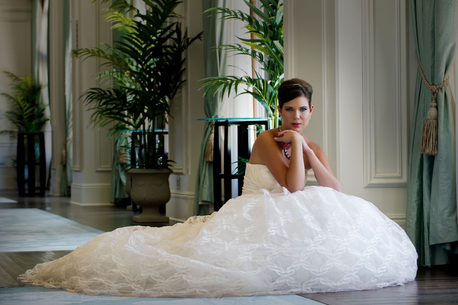 Elegant bride in a white ball gown sitting gracefully on the floor in a luxurious room with tropical plants.