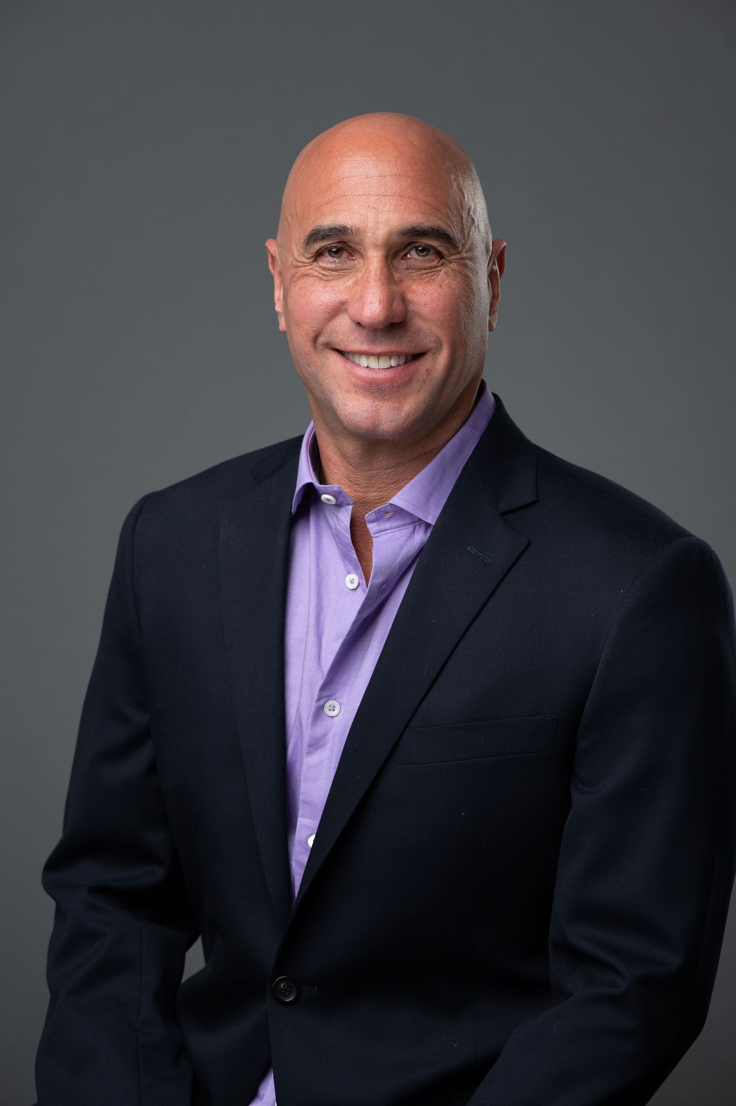 Professional headshot of a man in a navy blazer and purple shirt smiling confidently.