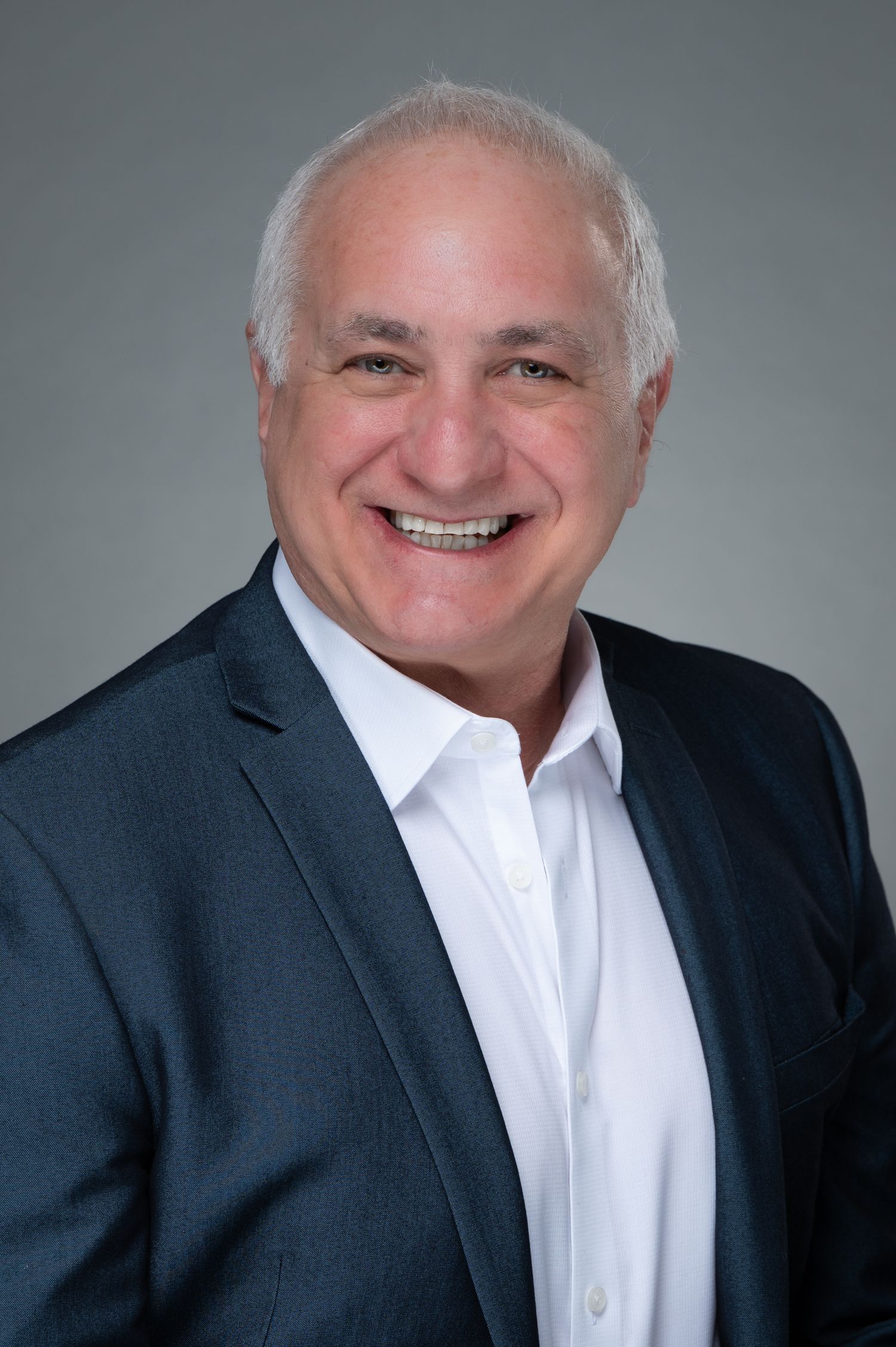 Smiling middle-aged man with short gray hair wearing a white shirt and dark blazer poses for a professional headshot.
