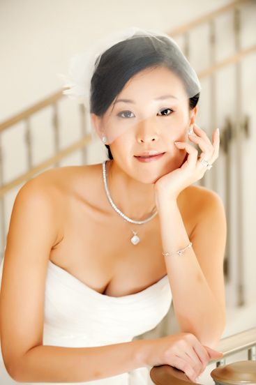 Elegant Asian bride in strapless white gown with veil, pearl necklace, and bracelet, posing gracefully on staircase.