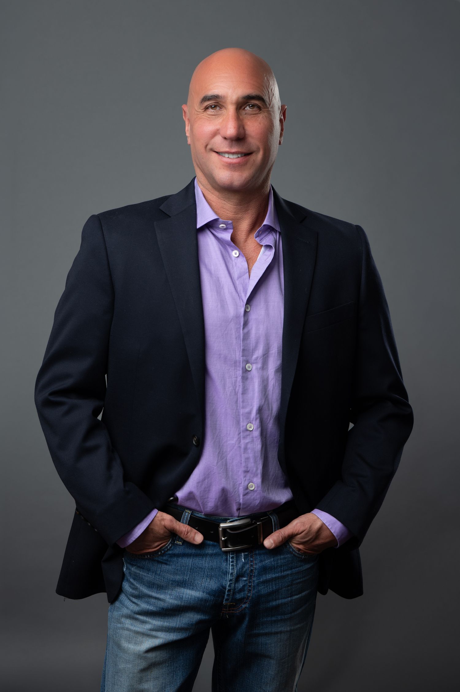 Confident bald man in purple shirt and navy blazer with jeans poses with hands in pockets against gray background.