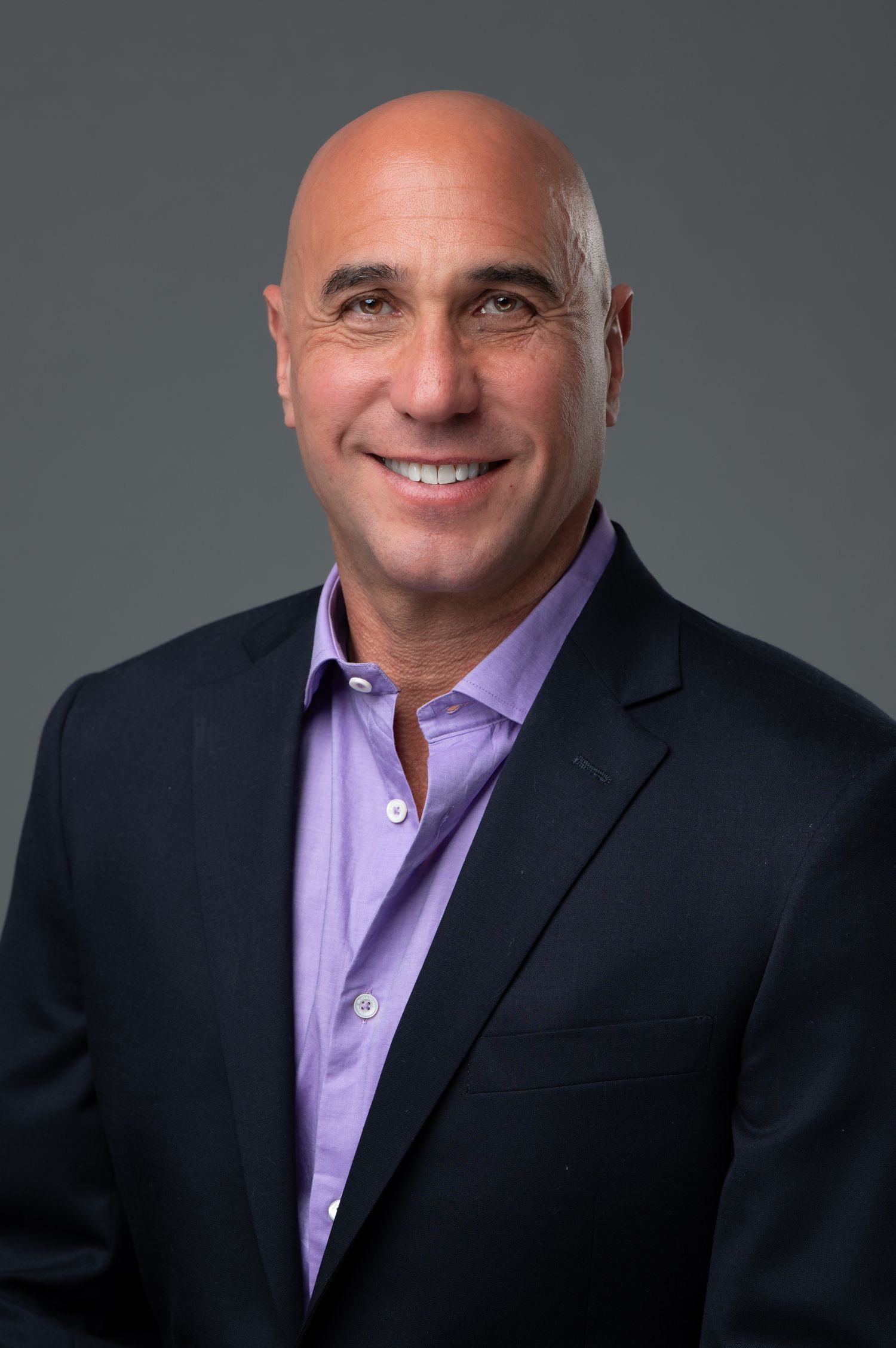 Smiling bald man in a purple shirt and dark blazer poses for a professional headshot against a gray background.
