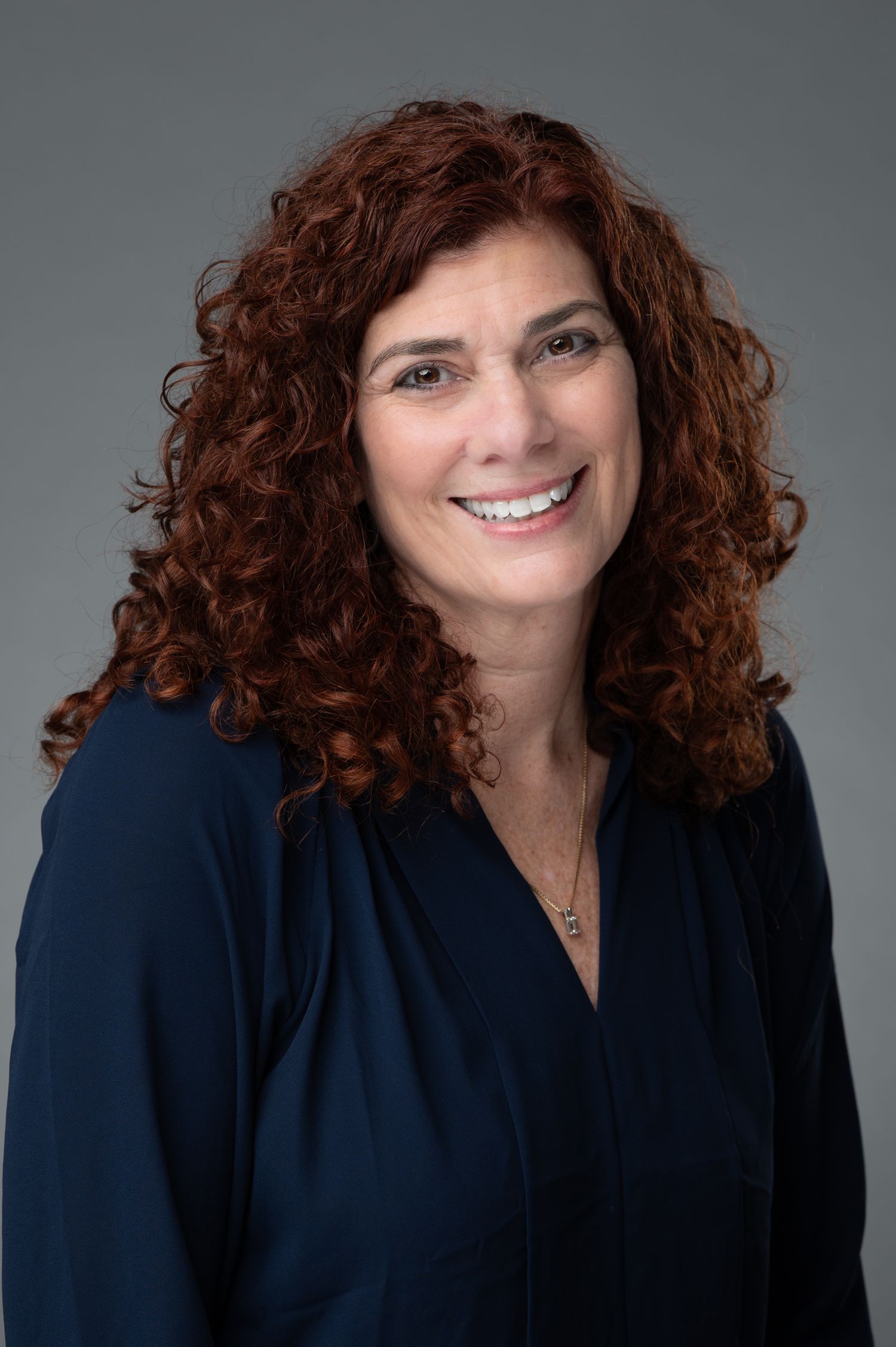 Professional headshot of a smiling woman with curly red hair wearing a navy blouse and delicate necklace.