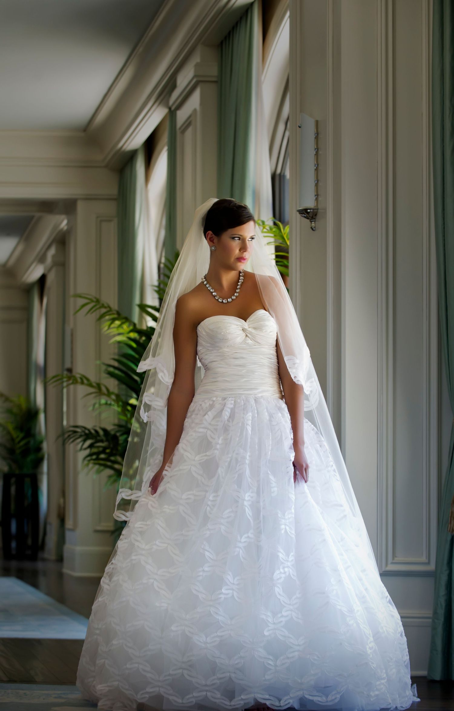 Bride in strapless white ball gown with lace, pearl necklace, and veil poses in elegant venue.