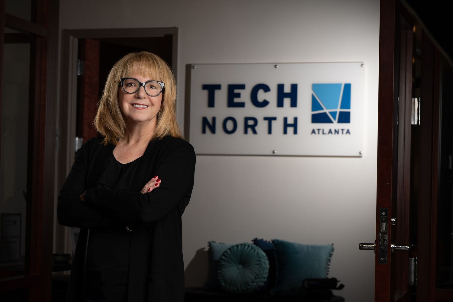 Woman with blonde hair and glasses smiling in front of Tech North Atlanta sign in modern office.