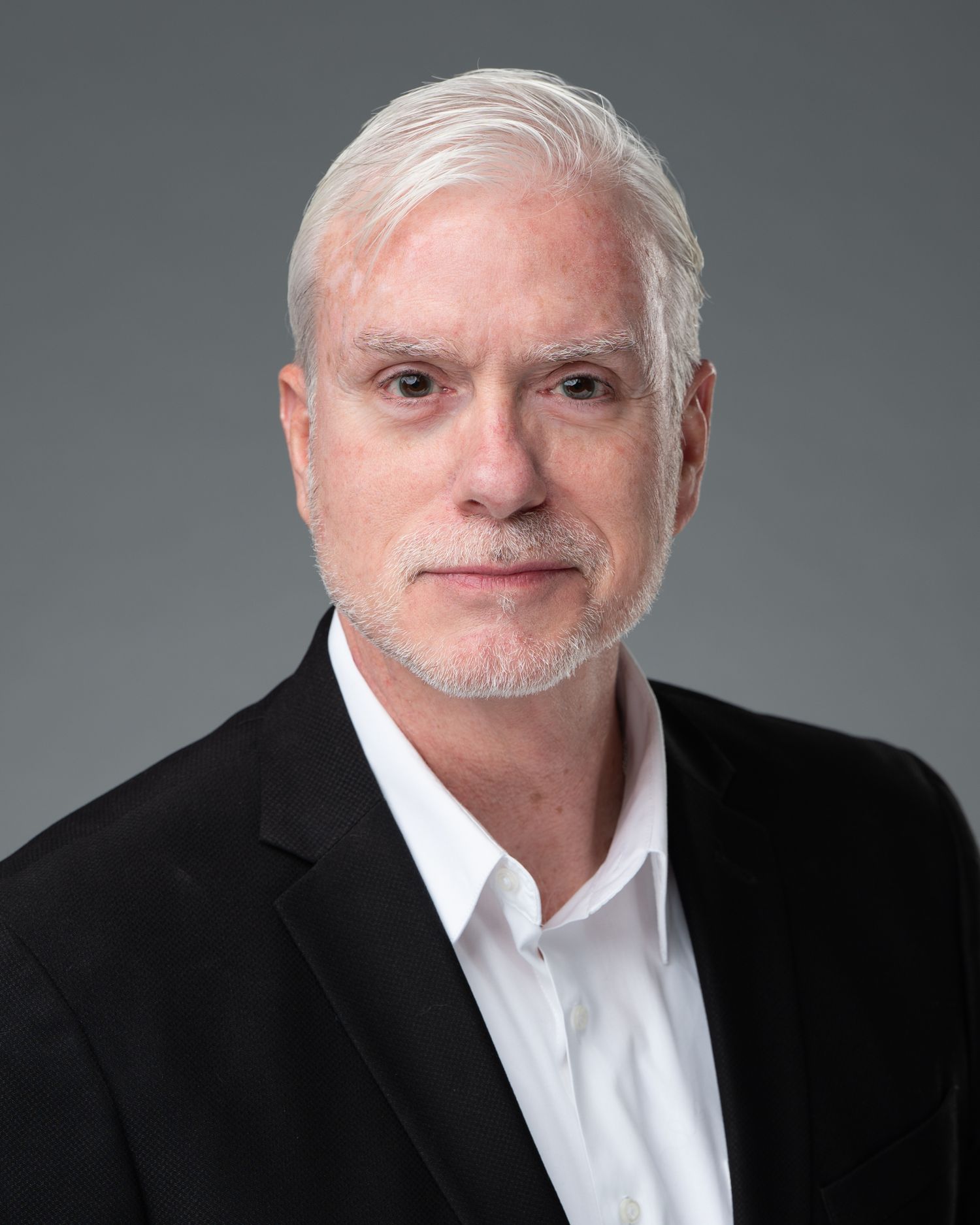 Professional headshot of a middle-aged man with white hair and beard wearing a black blazer and white shirt.