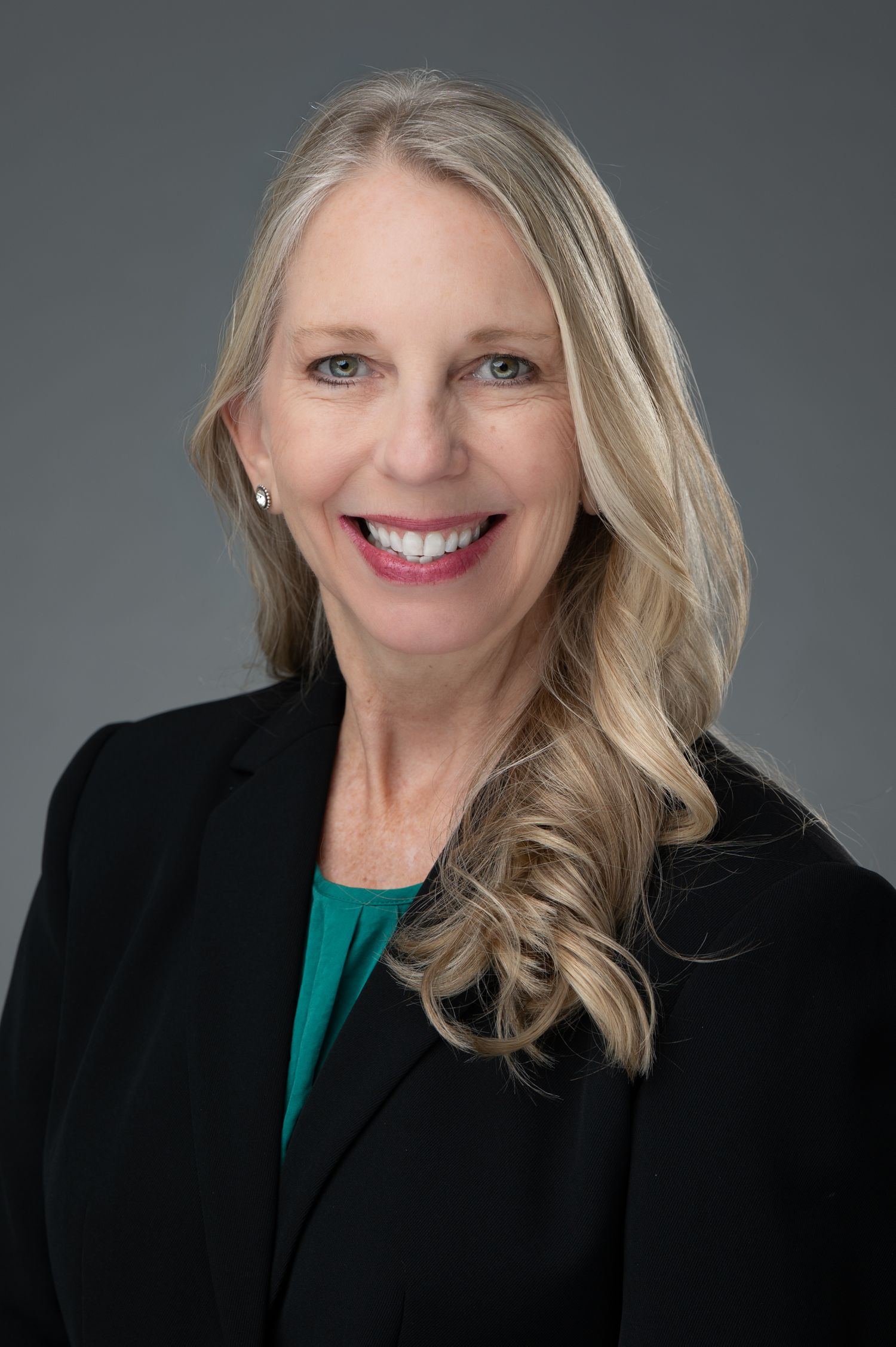 Professional headshot of a smiling woman with blonde hair wearing a black blazer and teal top against a gray background.