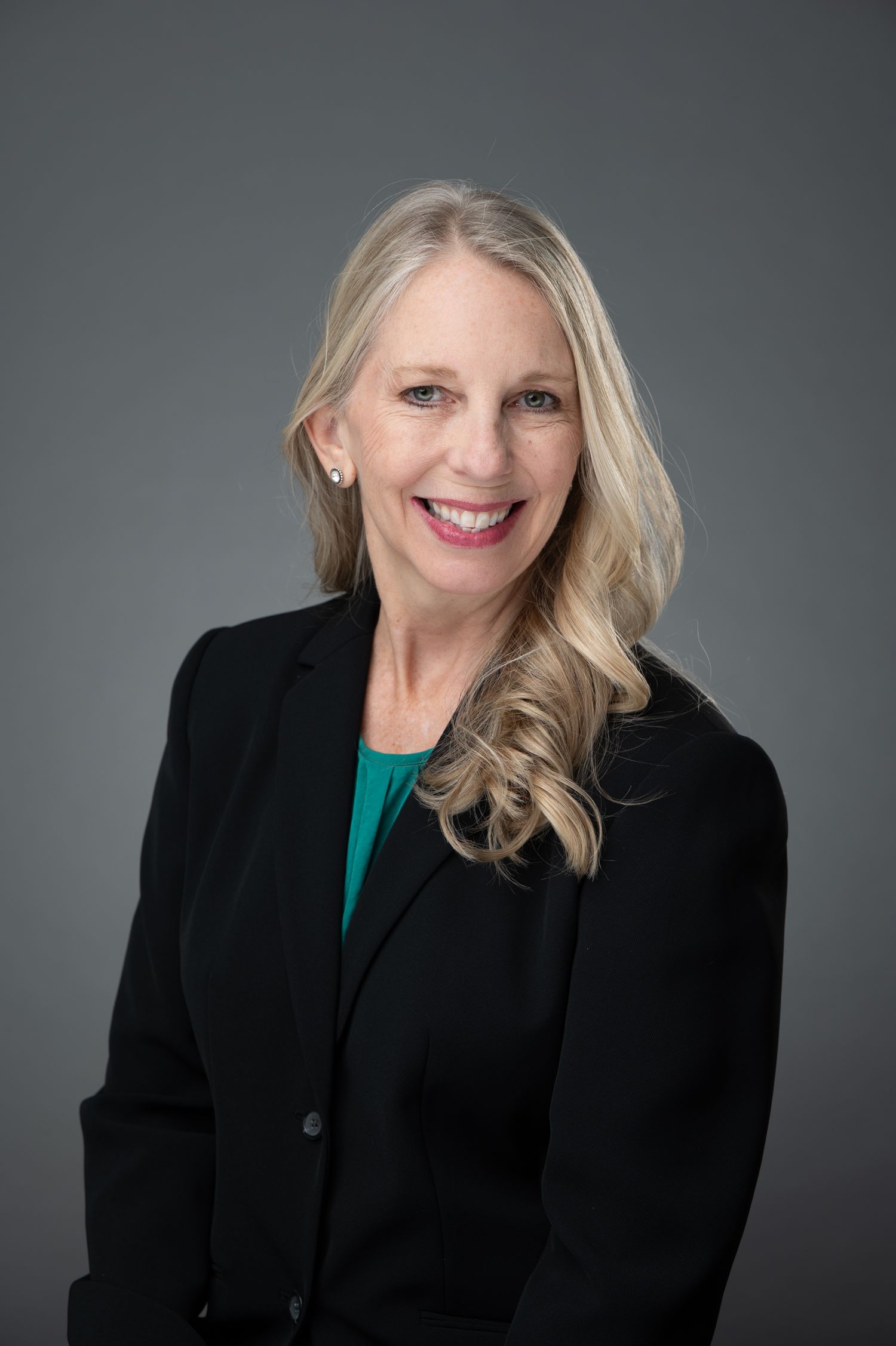 Professional headshot of a smiling blonde woman in a black blazer and teal top against a gray background.