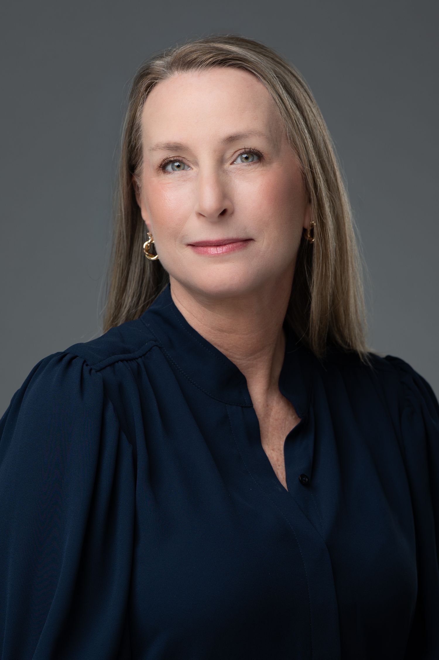 Professional headshot of a woman with long blonde hair wearing a navy blouse and gold earrings against a gray background.
