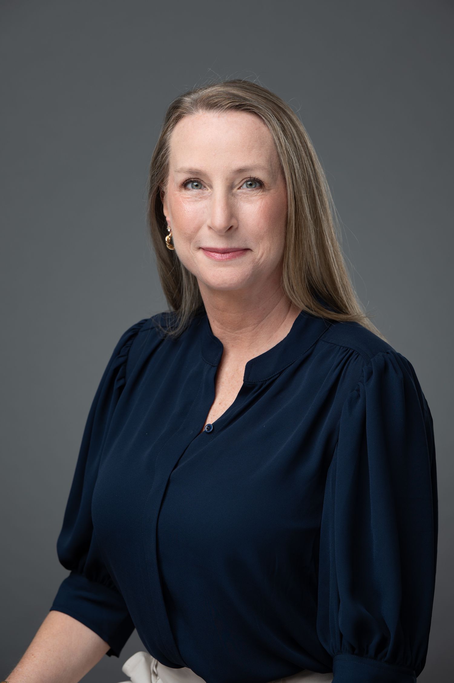 Professional headshot of a woman with long blonde hair wearing a navy blue blouse against a gray background.