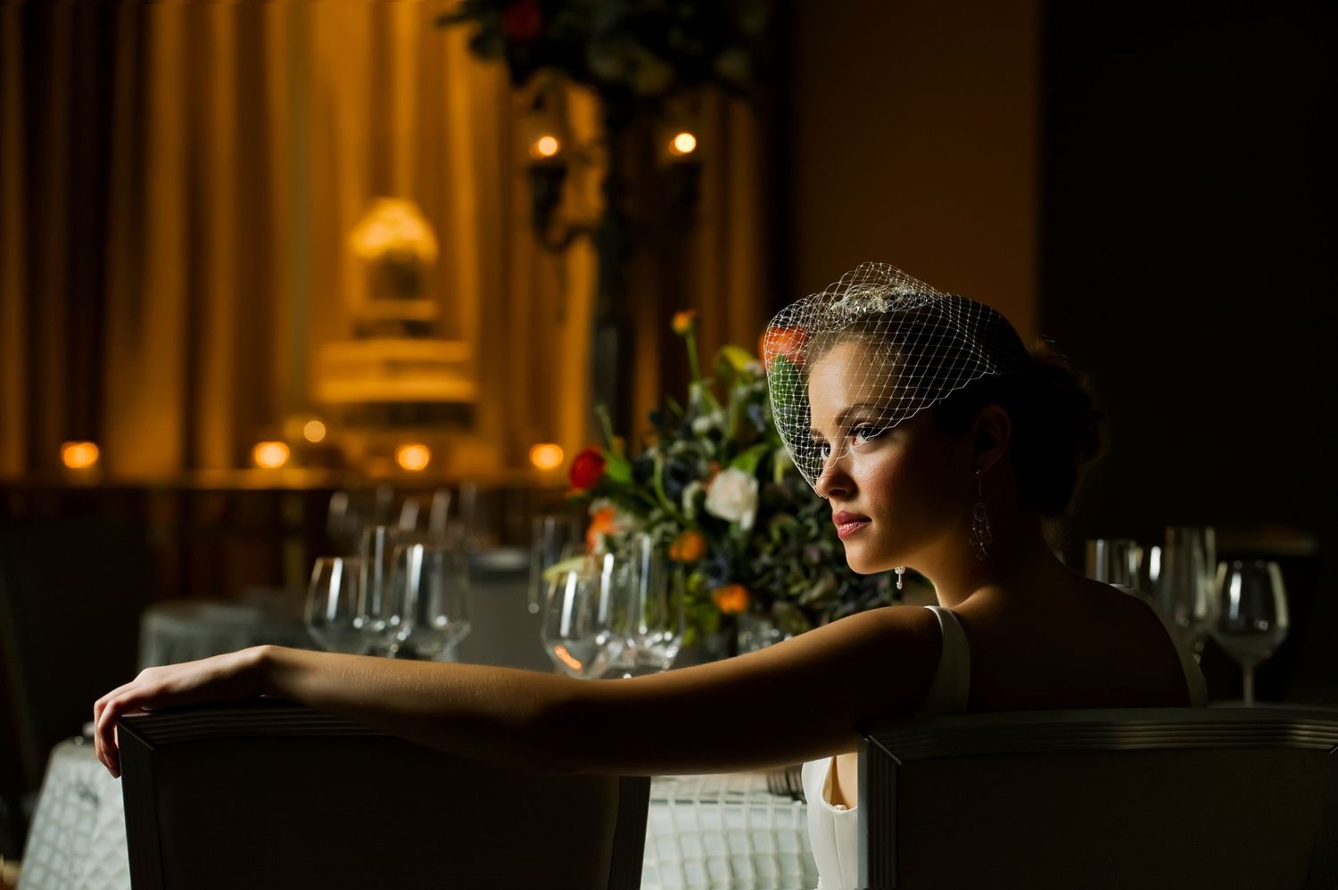 Elegant bride wearing a birdcage veil seated at a decorated wedding reception table with candles and floral arrangements.