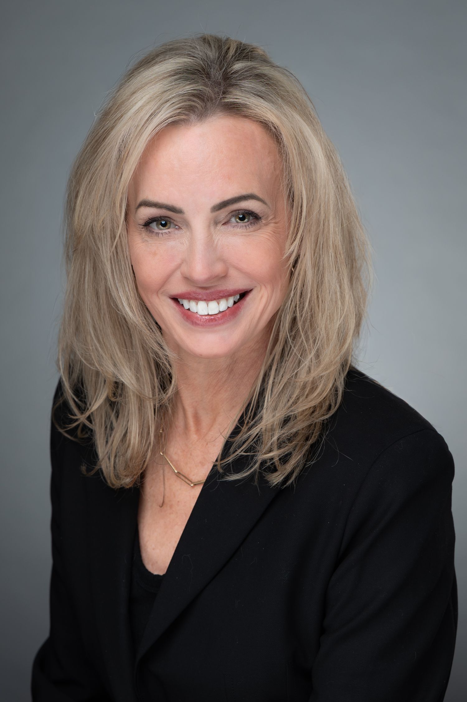Professional headshot of a blonde woman in a black blazer smiling against a gray background.