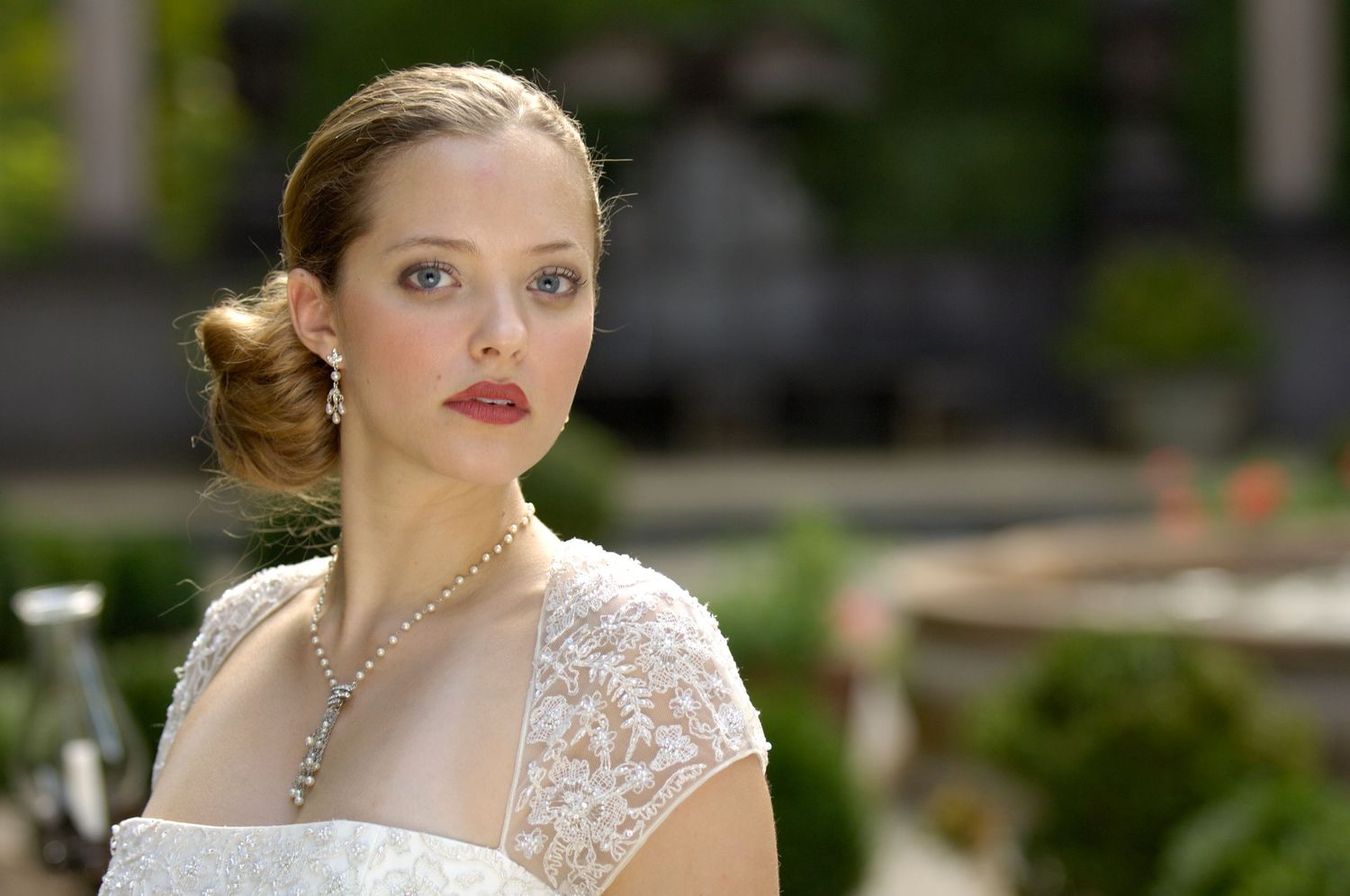 Elegant bride in white lace dress with pearl necklace and updo hairstyle posing outdoors.