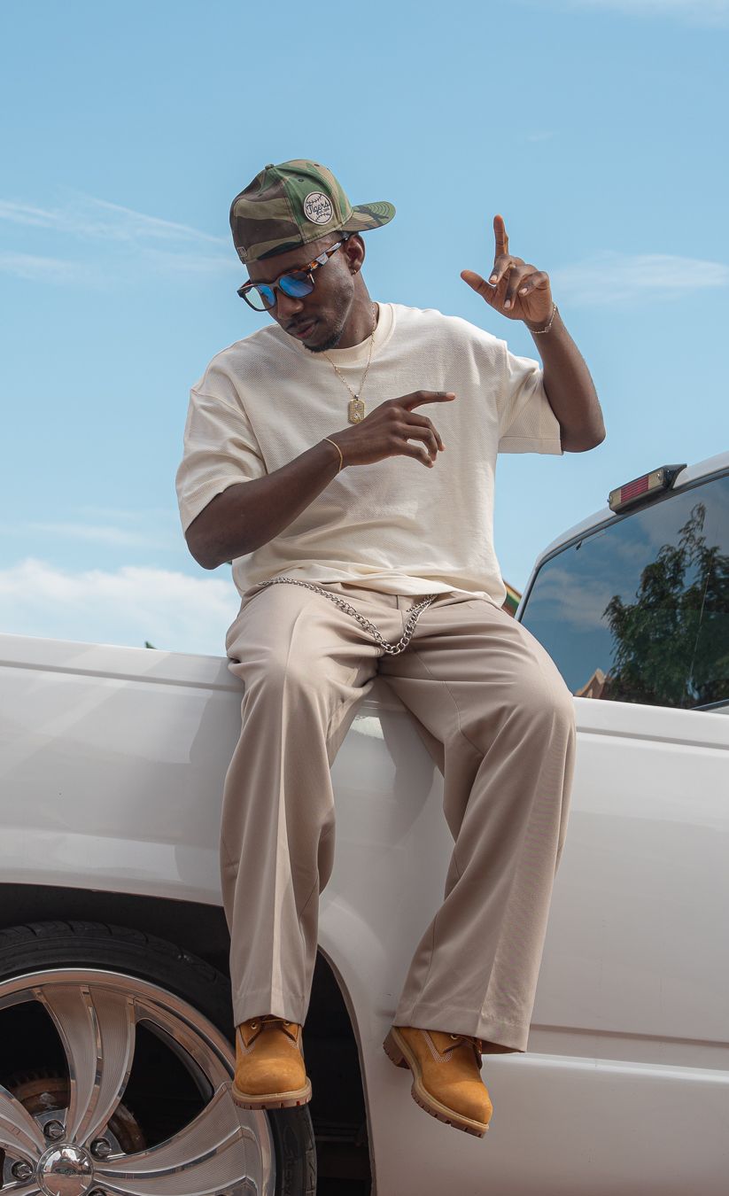 Young man in camo cap, cream outfit, and tan boots posing on white car hood under blue sky.