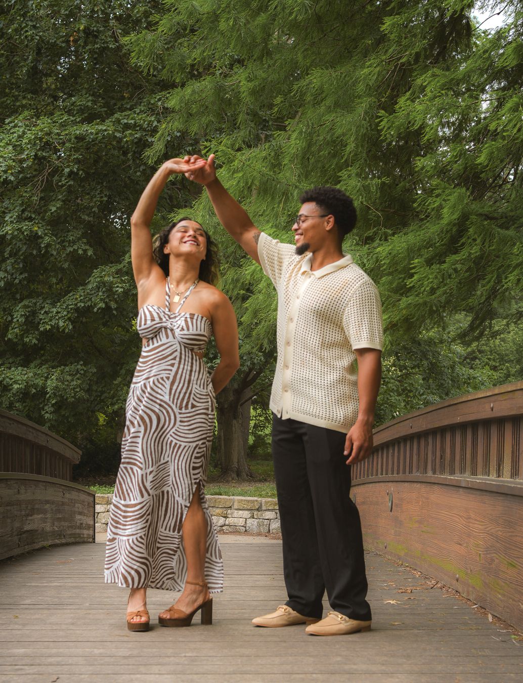 A couple dances gracefully on a wooden bridge surrounded by lush green trees, the woman in a patterned dress.