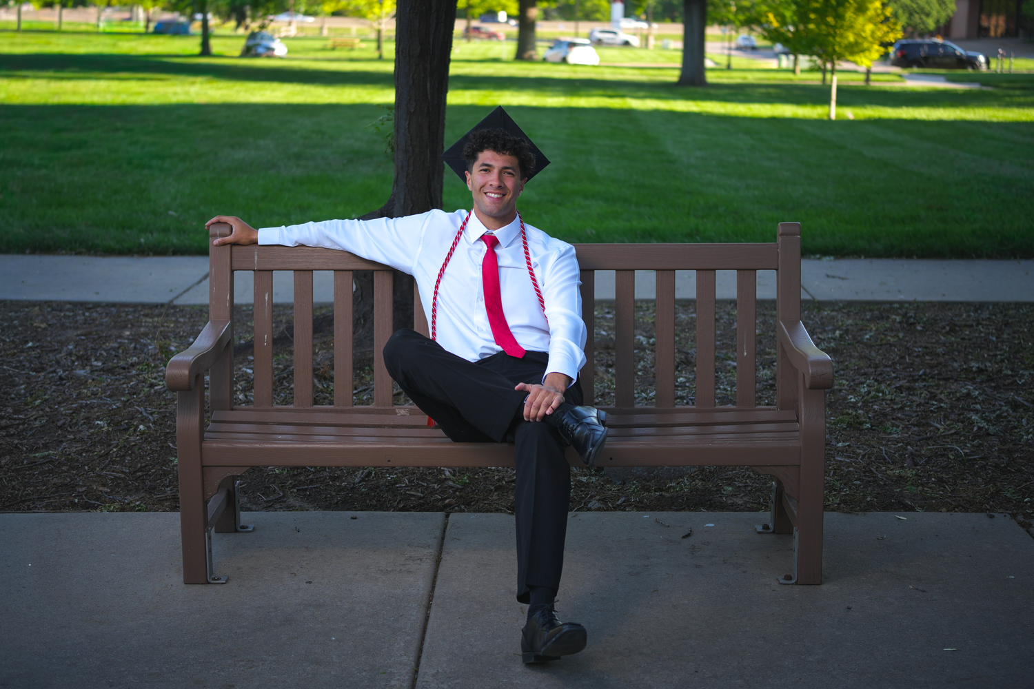 Graduate in cap and red suspenders relaxes on wooden bench on a sunny college campus.