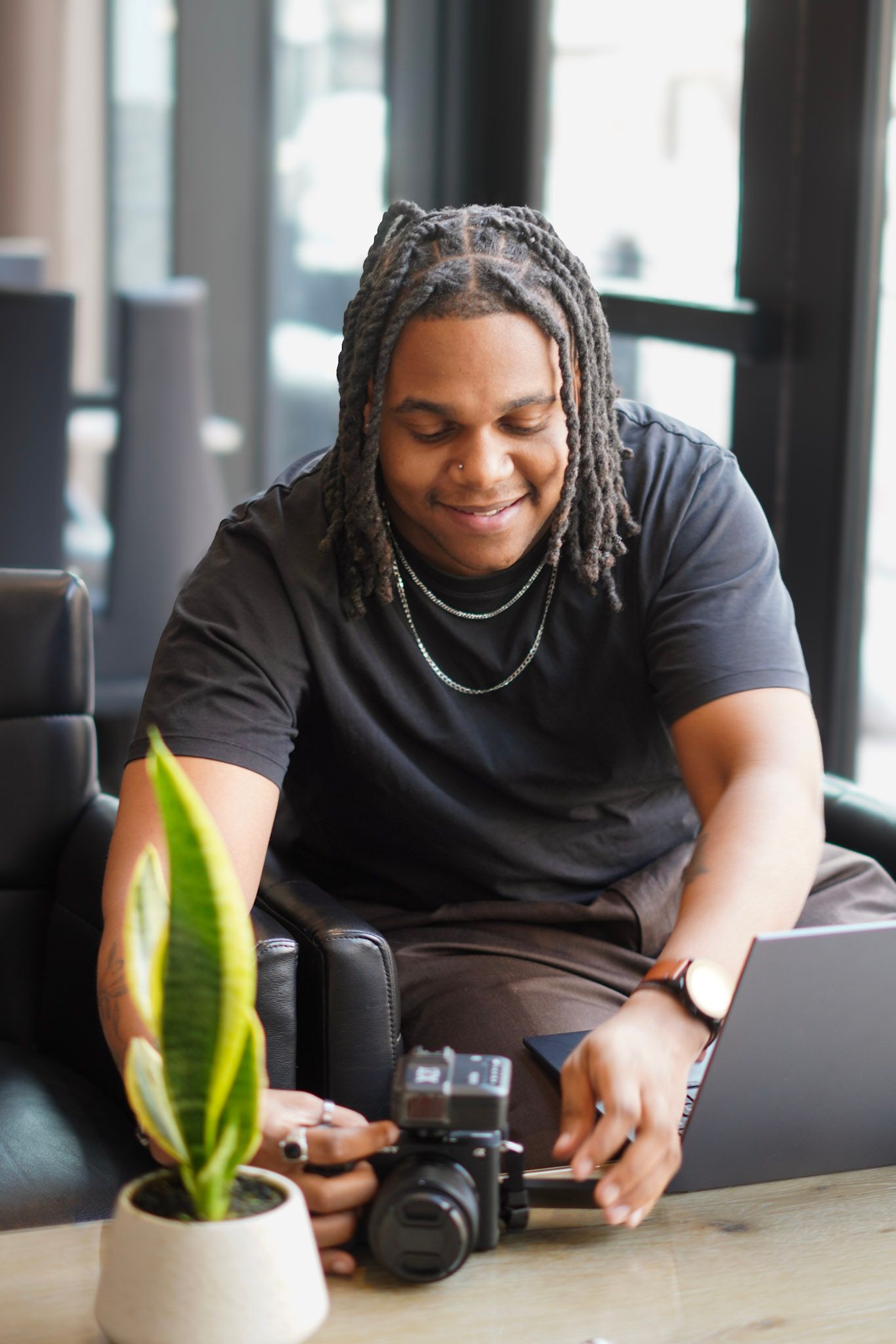 A smiling young Black man with braids works on a laptop beside a Sony camera at a table with a plant.