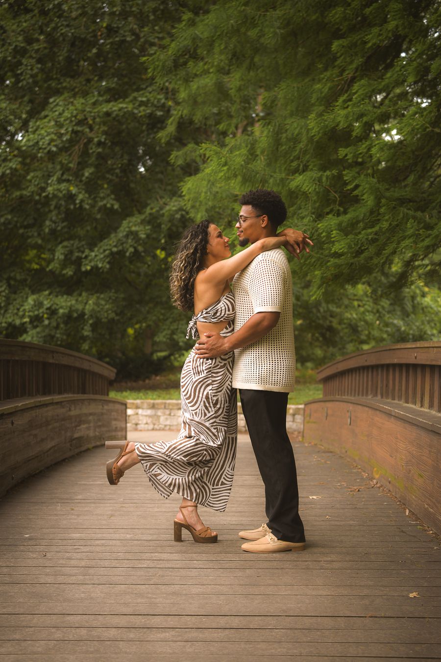 Couple embracing and dancing on a wooden bridge surrounded by lush green trees in a romantic outdoor setting.