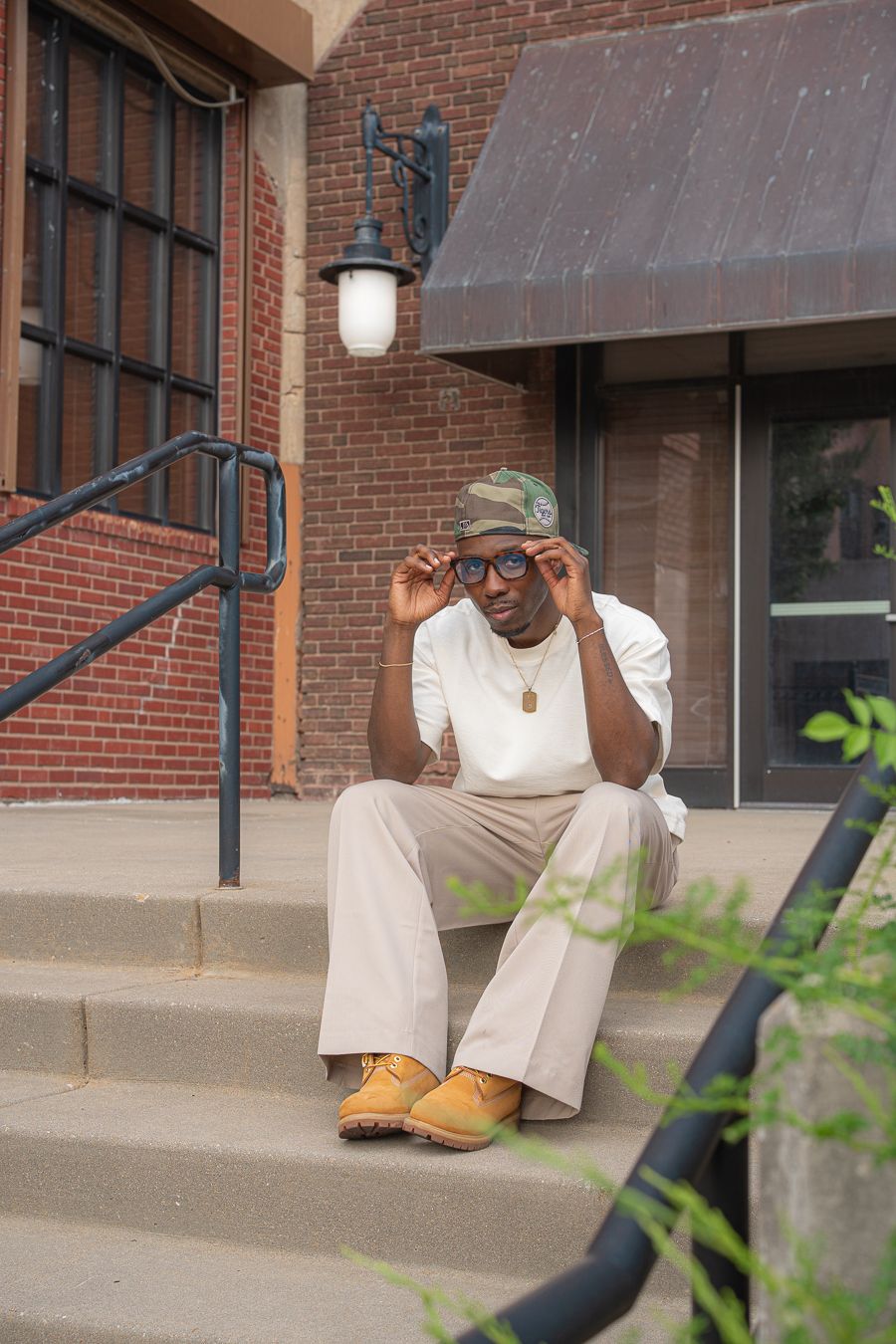 Man in camo cap, glasses, white tee, beige pants, and tan boots sitting on urban building steps.