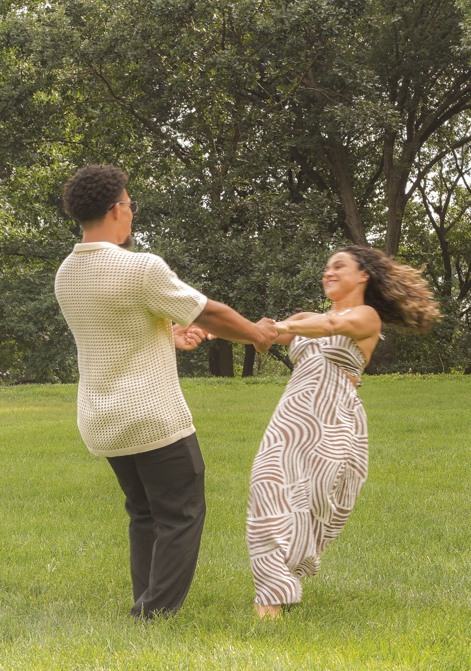 A young couple dances joyfully in a lush green park, holding hands and spinning together outdoors.