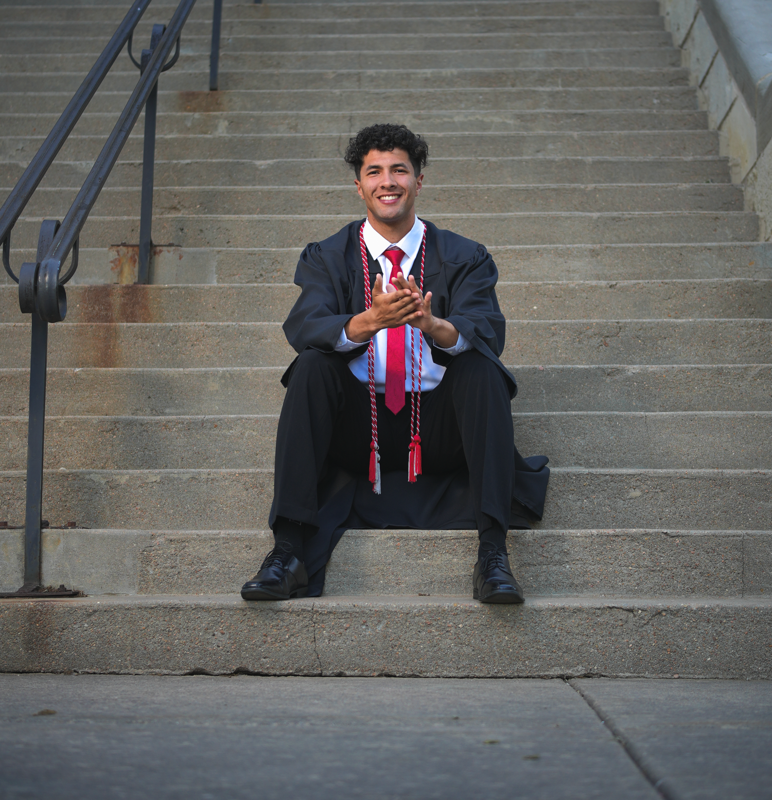 Smiling male graduate in black gown and red tie sits on outdoor concrete steps clapping hands.