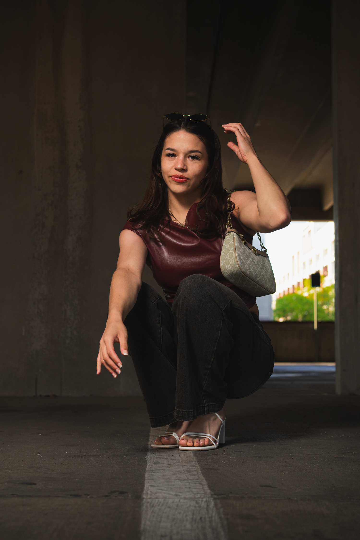 Young woman in burgundy leather crop top crouches under concrete overpass holding designer bag.