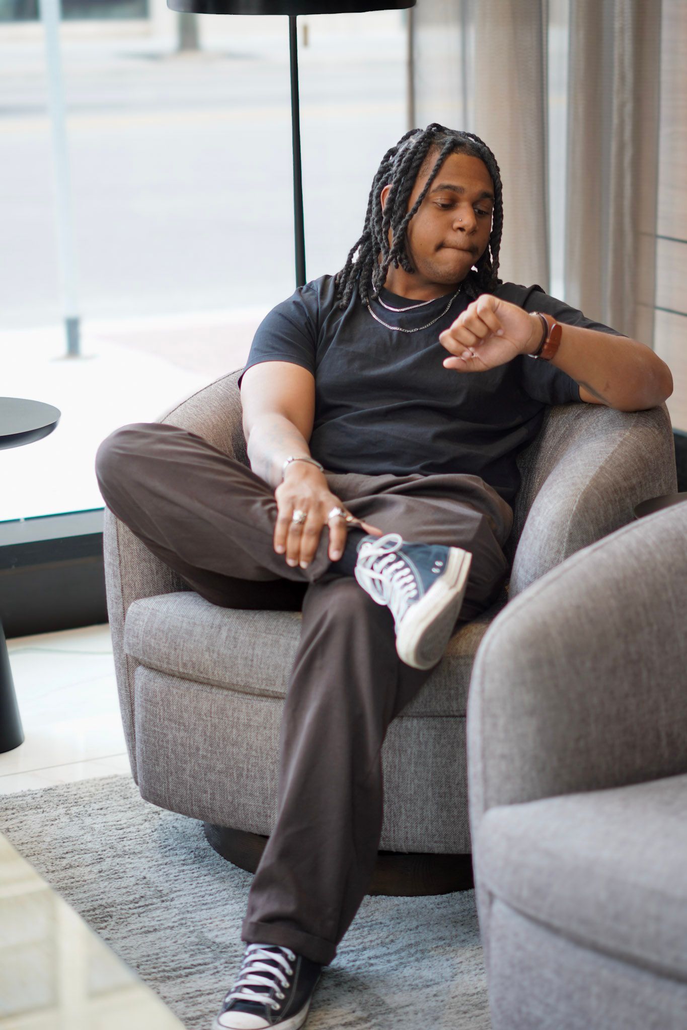 A young Black man with braids checks his watch while seated in a gray armchair in a modern hotel lobby.