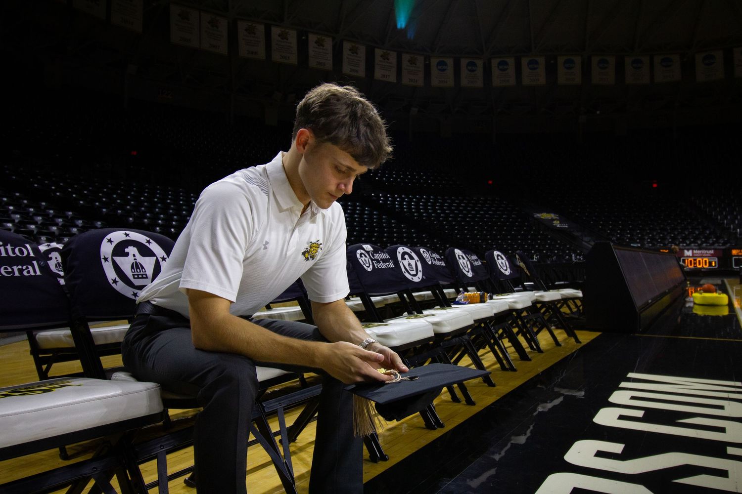 Graduate in white polo sits focused at a table on a basketball court in an arena.